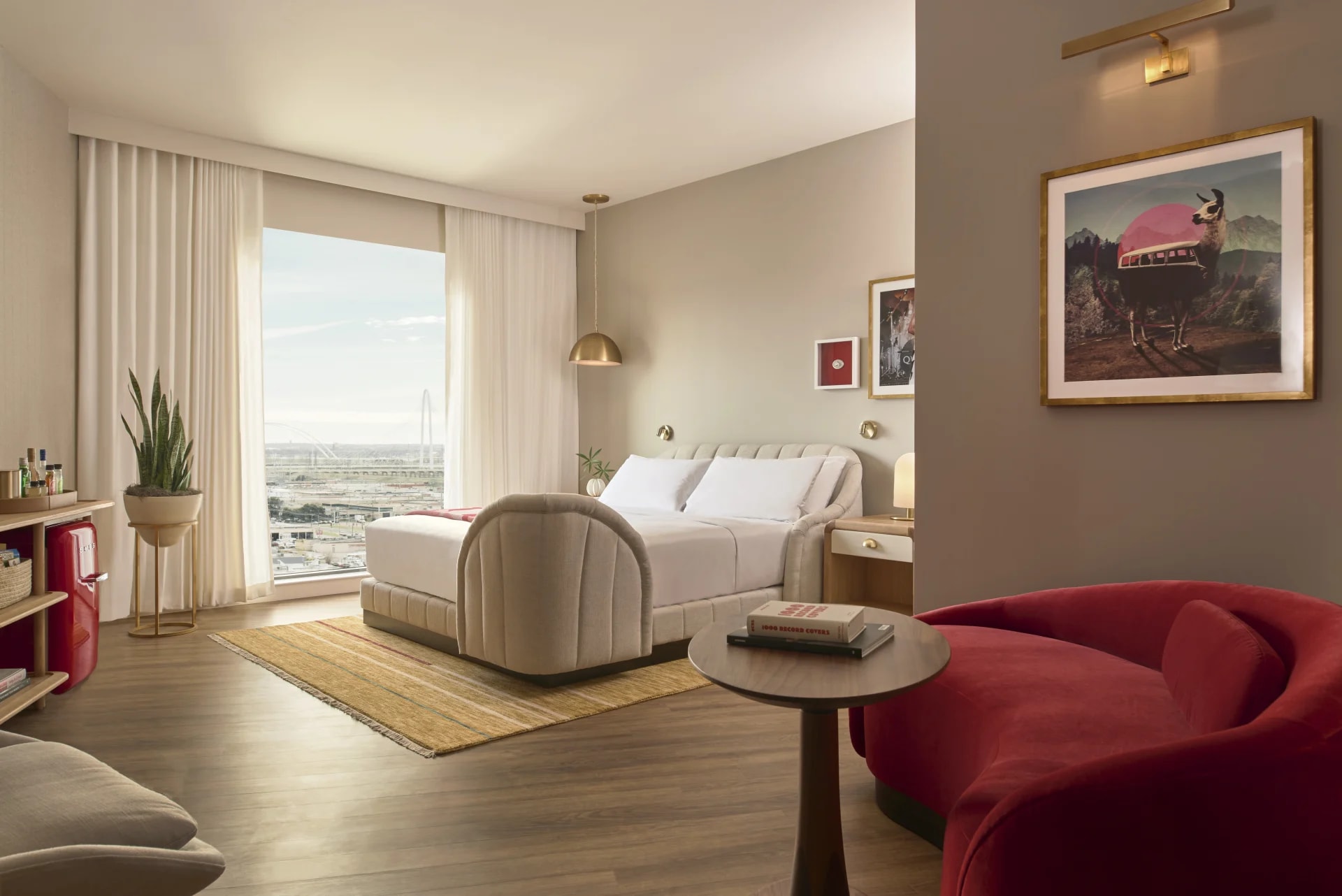 Stylish hotel room with neutral tones, a plush bed, and a red velvet sofa, featuring mid-century decor and a large window overlooking the city skyline.