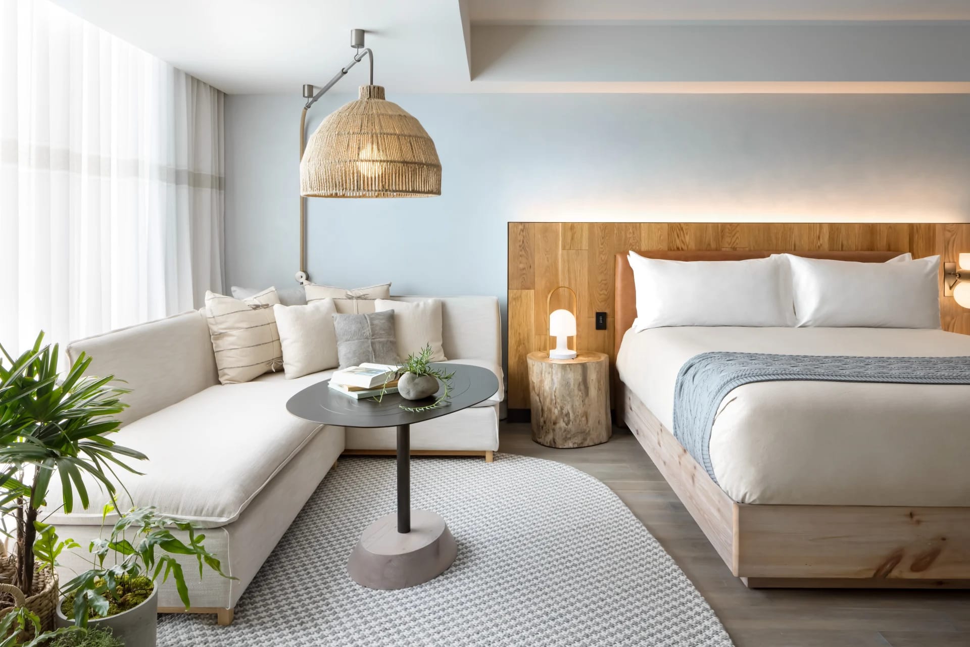 Modern hotel room with natural wood accents, neutral tones, and soft textures, featuring a cozy bed, sectional sofa, woven pendant light, and potted plants adding a touch of greenery.