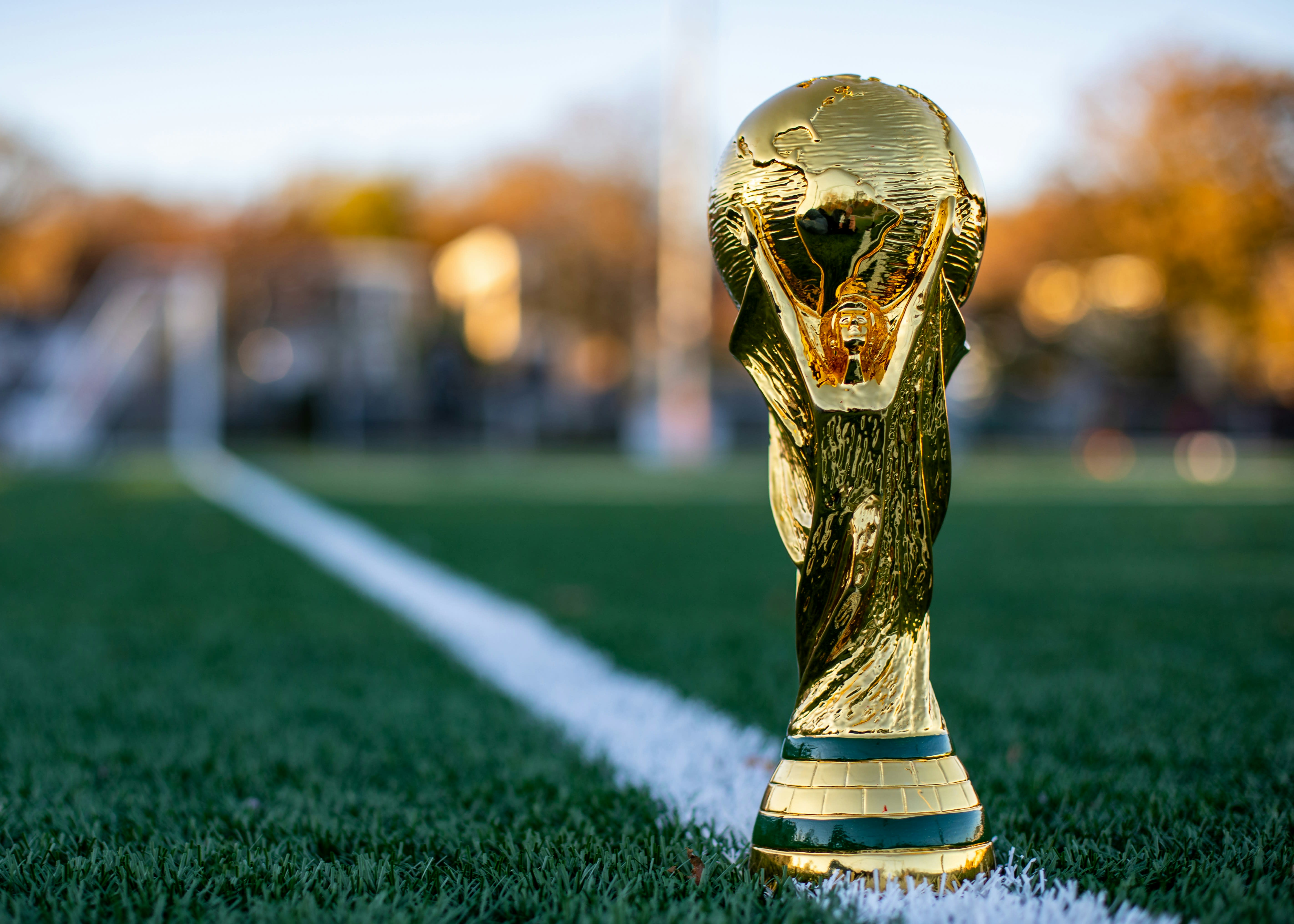 The FIFA World Cup trophy placed on a green soccer field, gleaming in the sunlight with a blurred background of trees and stadium surroundings.