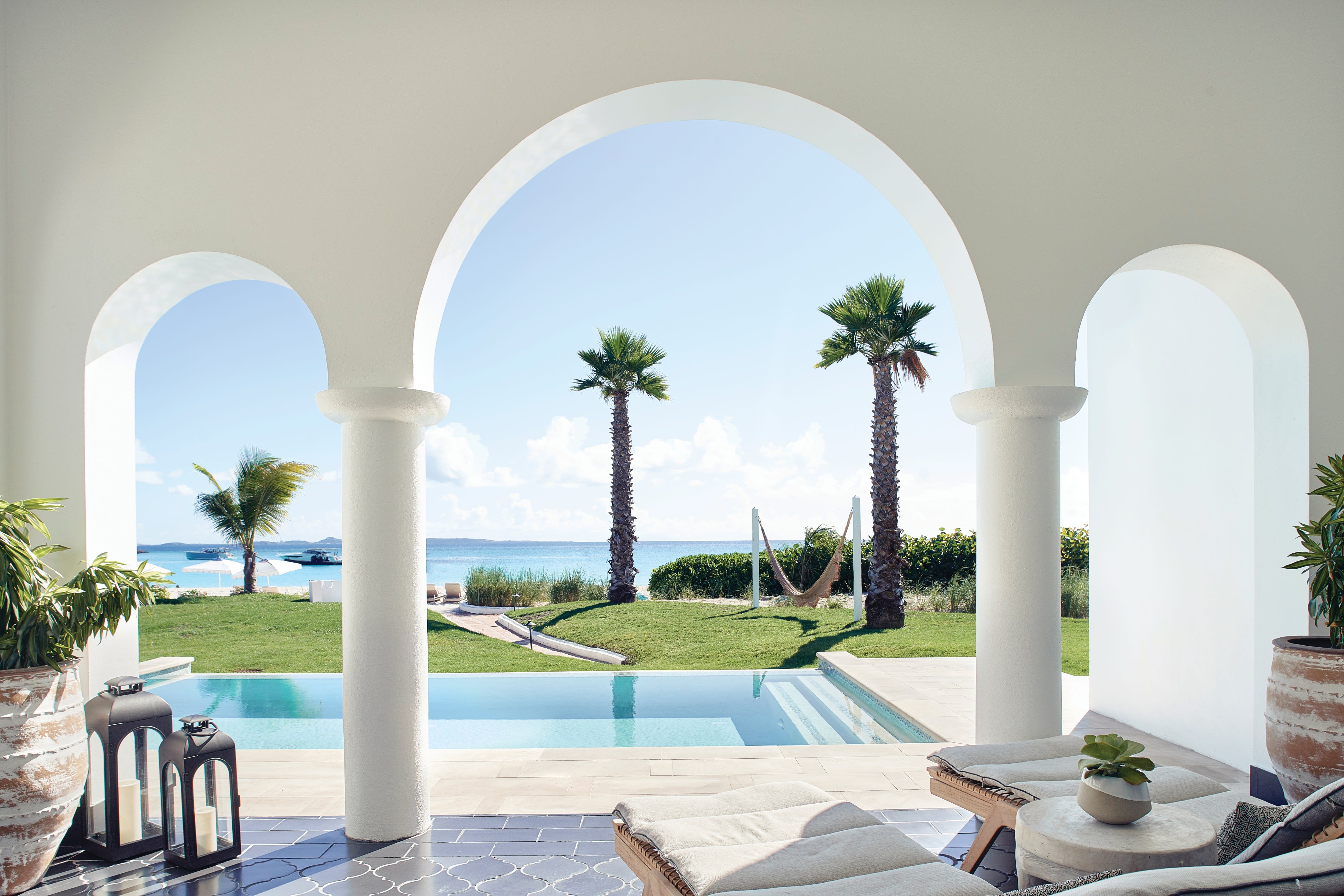 Serene oceanfront villa with white arches framing views of palm trees, a private pool, and a hammock overlooking the turquoise sea under a clear blue sky.