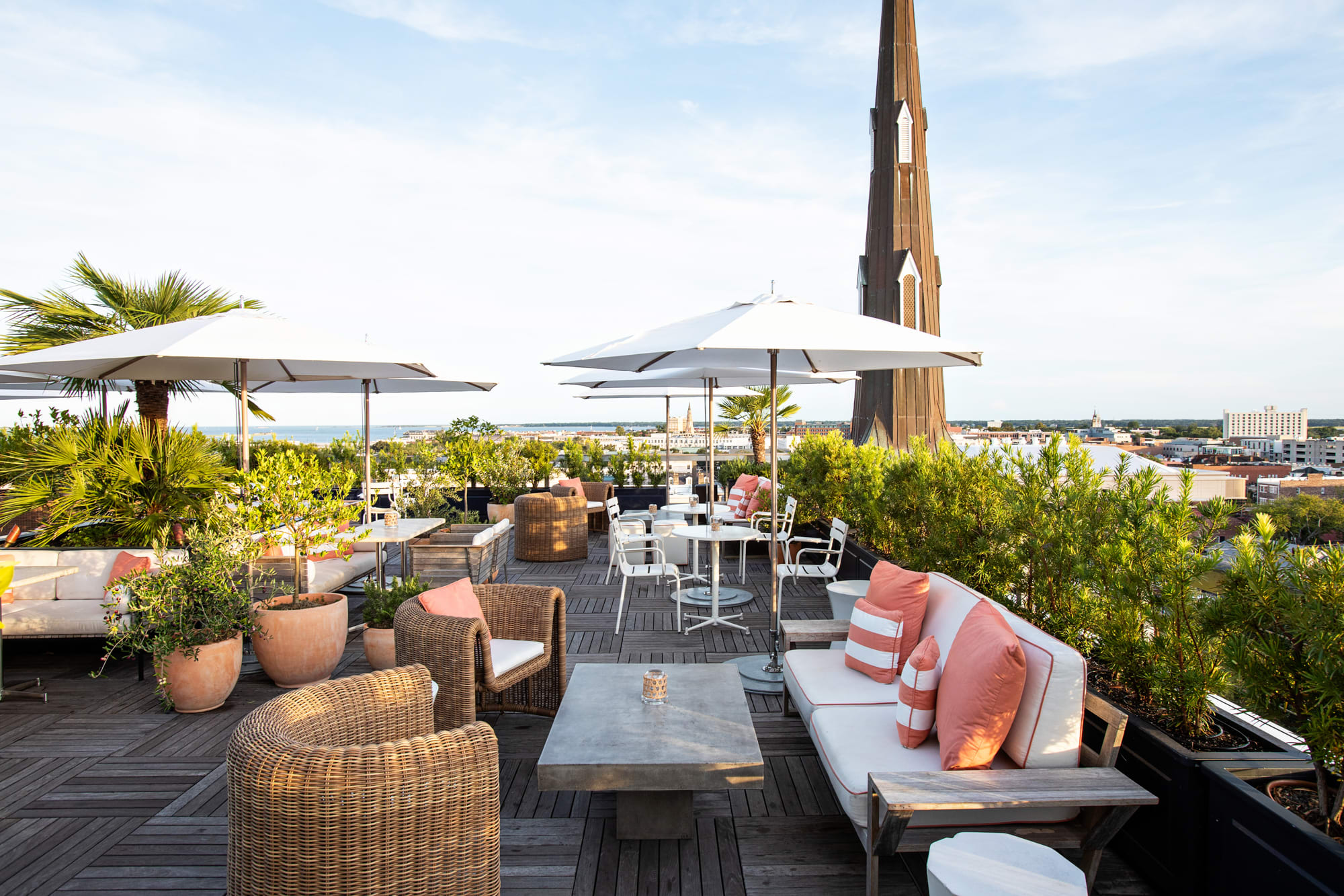 Rooftop terrace bar with cushioned seating, coral-striped pillows, white umbrellas, and city views framed by palm trees and a tall church steeple in the background.