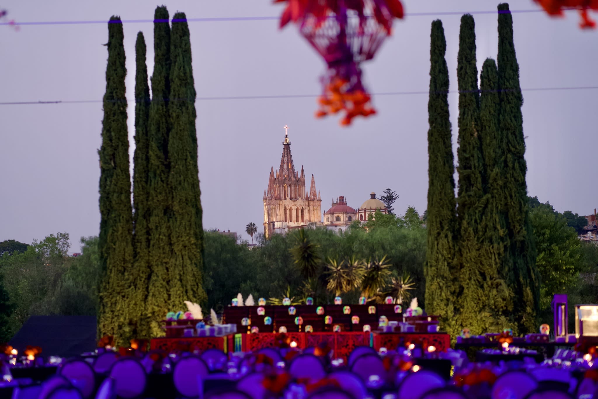 Evening view of the Parroquia de San Miguel Arcángel framed by tall cypress trees, with colorful Day of the Dead decorations and glowing candles in the foreground.