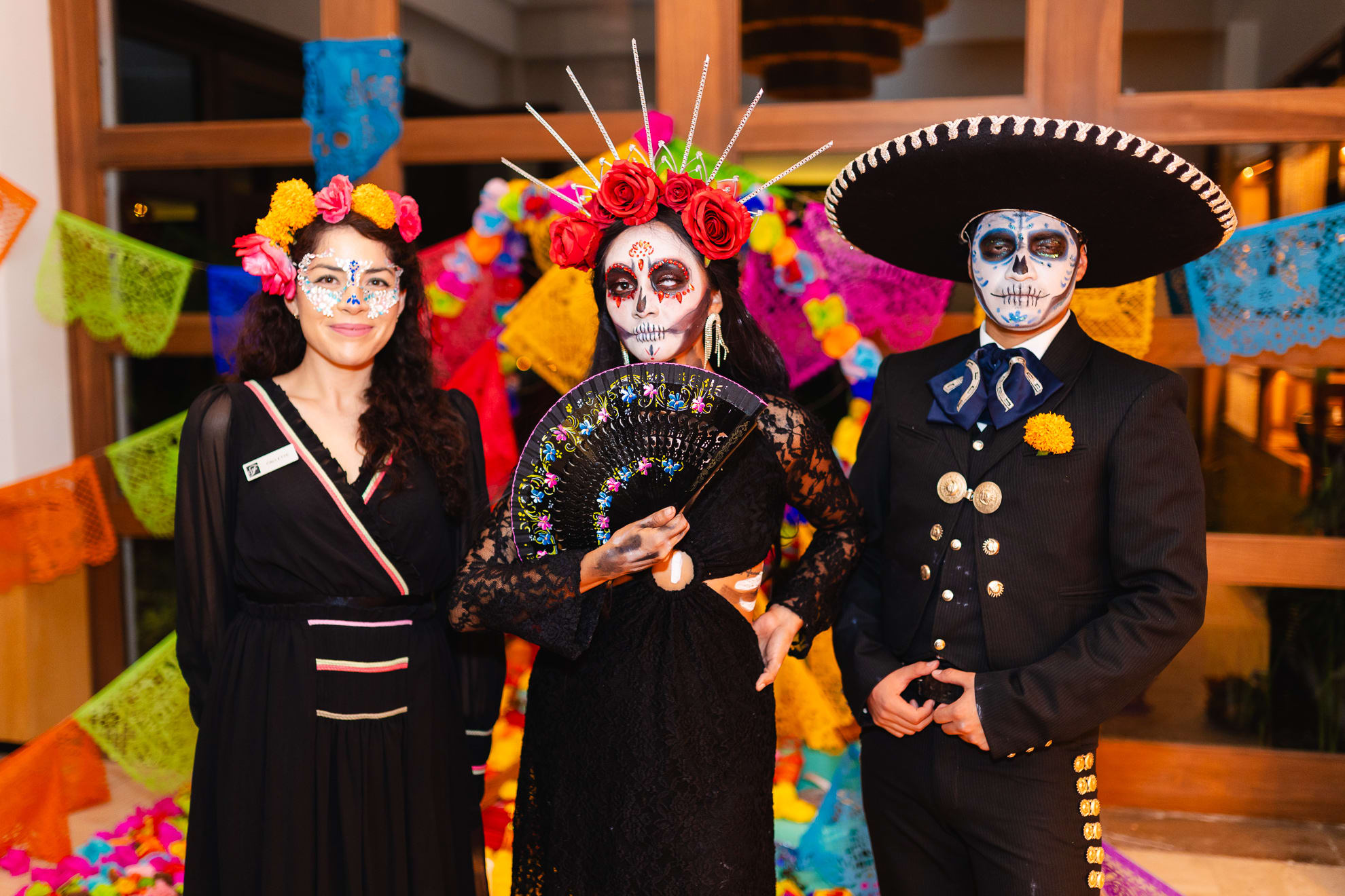Three people dressed in elegant Día de Muertos attire with painted calavera faces, wearing black clothing adorned with flowers and traditional accessories, standing before colorful papel picado decorations.
