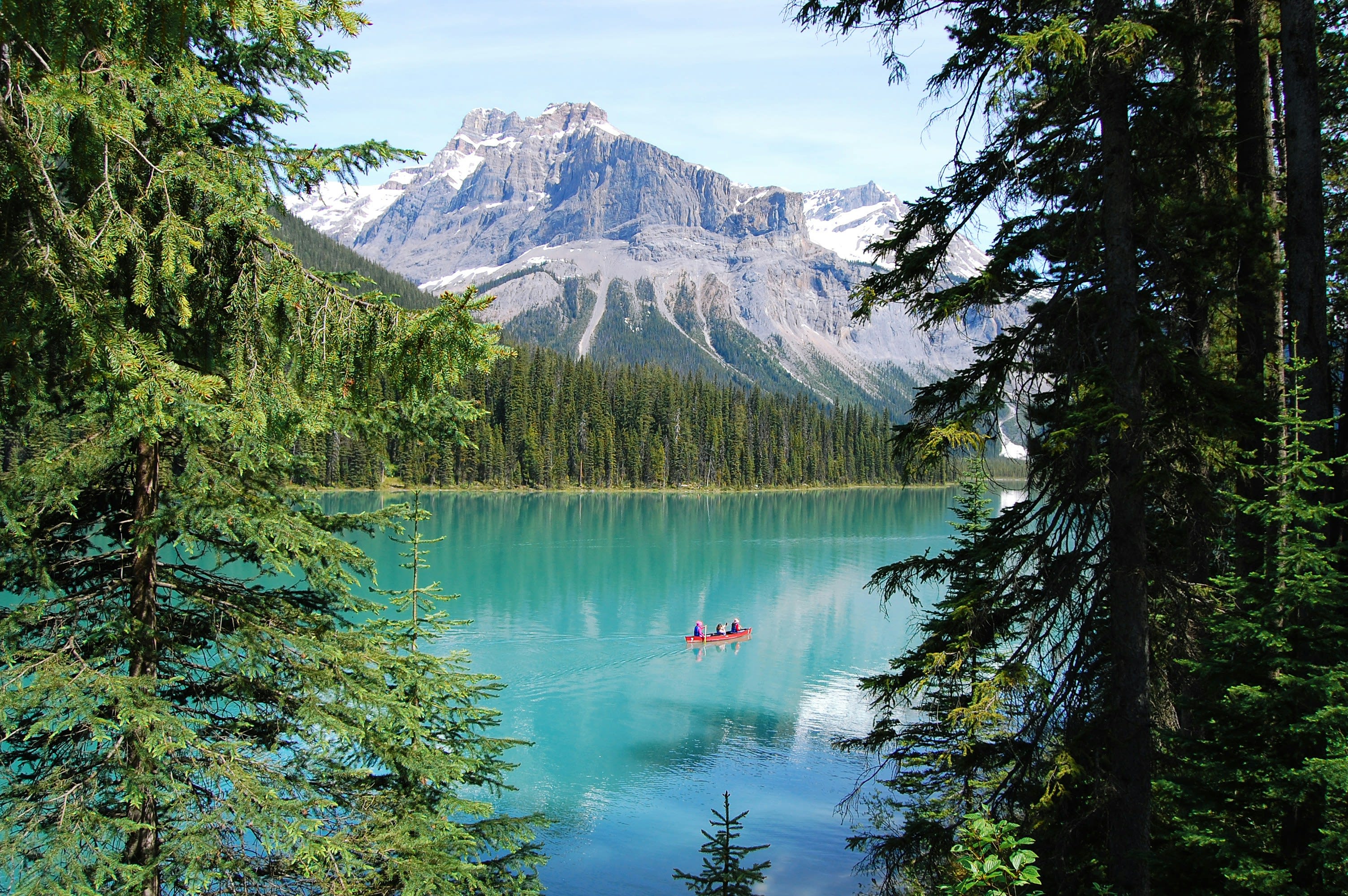 Turquoise lake surrounded by dense evergreen forest and snow-capped mountains, with a red canoe carrying people gliding across the calm water under a clear blue sky.