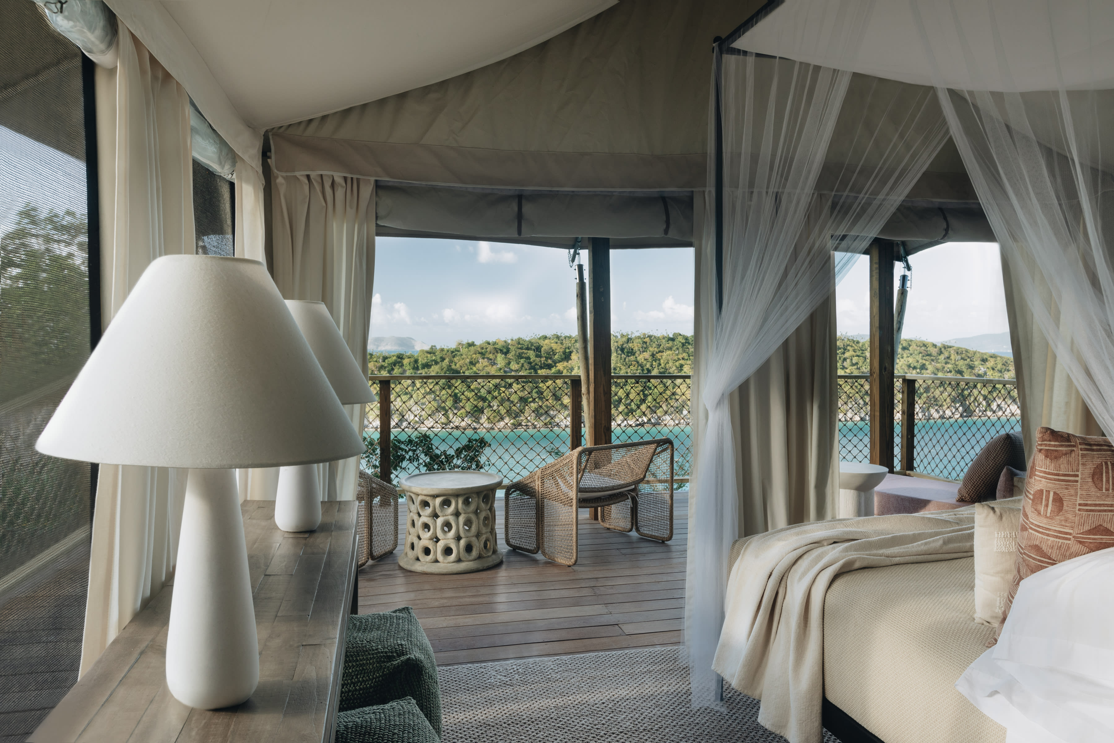 Elegant glamping tent bedroom with neutral tones, canopy bed, and open terrace overlooking turquoise water and lush green hills.
