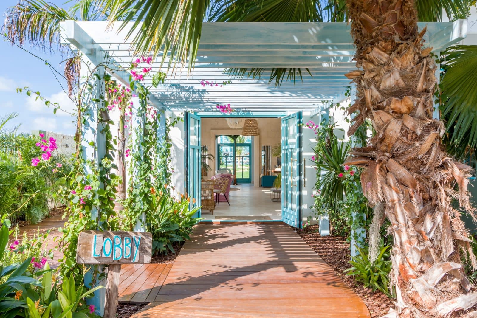 Bright tropical hotel entrance framed by palm trees and blooming bougainvillea, leading through turquoise double doors into a breezy, sunlit lobby.