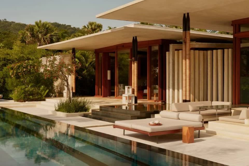 Modern villa with minimalist architecture, open glass walls, and a sleek pool surrounded by lush greenery and stylish sun loungers.