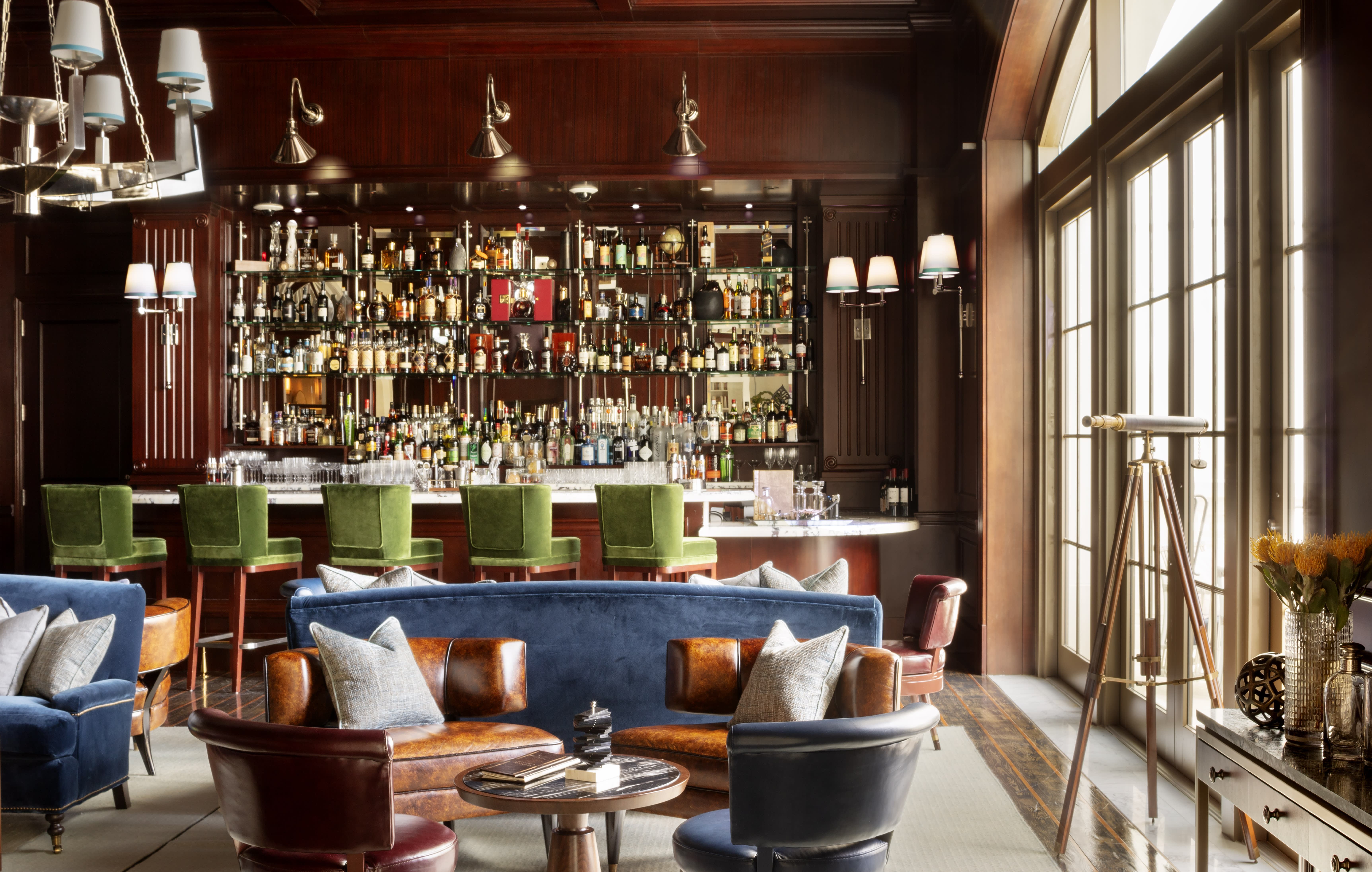 Sophisticated hotel bar with rich wood paneling, a mirrored back bar stocked with bottles, green velvet barstools, and plush leather and velvet seating in jewel tones.