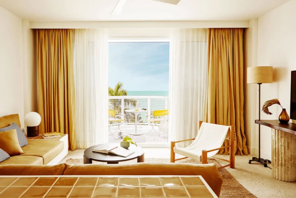 Bright, contemporary hotel room with golden curtains, a cozy seating area, and a balcony overlooking turquoise ocean views with palm trees and a sunny blue sky.