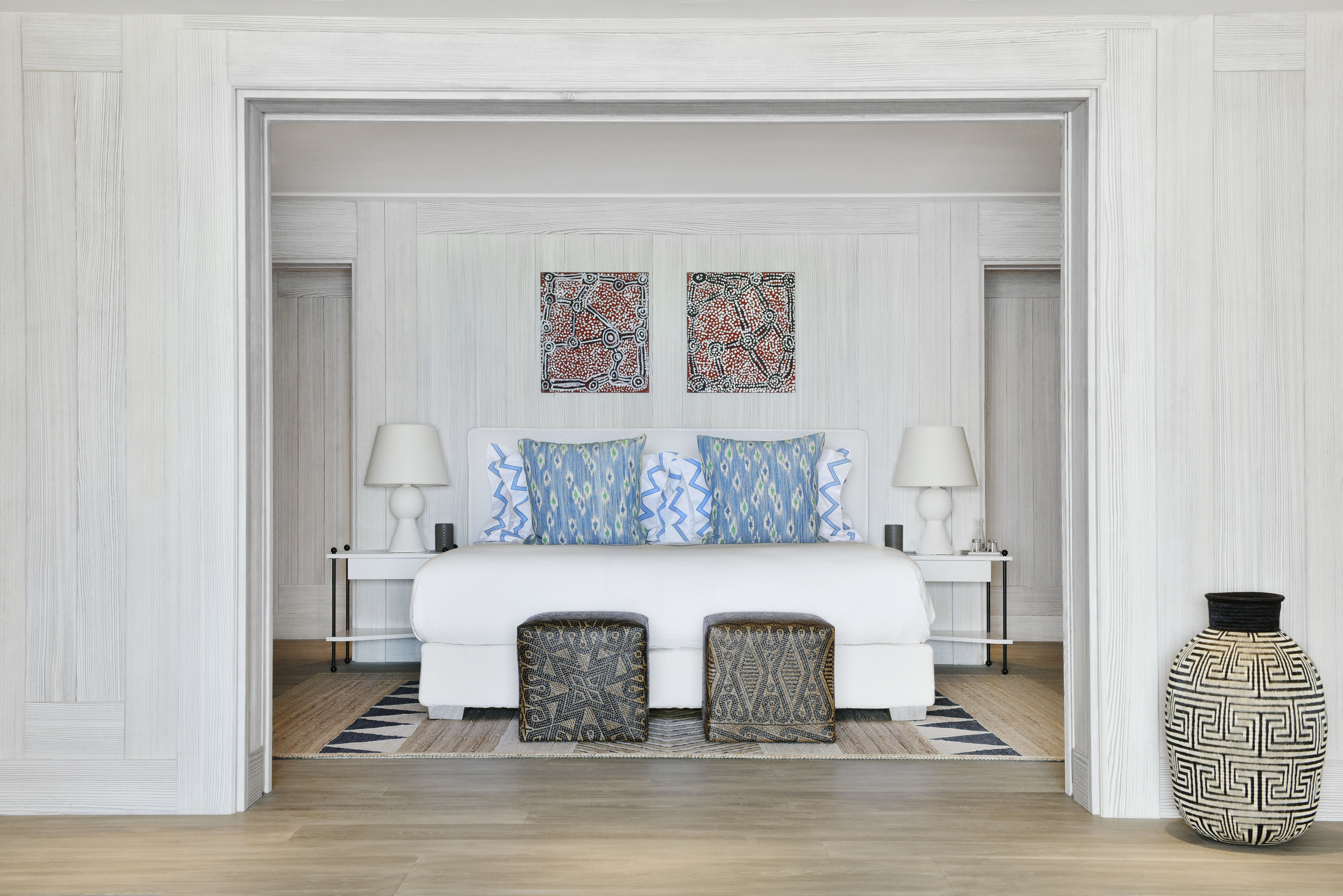 Elegant hotel bedroom with whitewashed wood walls, a king bed accented by blue patterned pillows, tribal artwork above the headboard, and woven stools and decor adding a touch of island style.