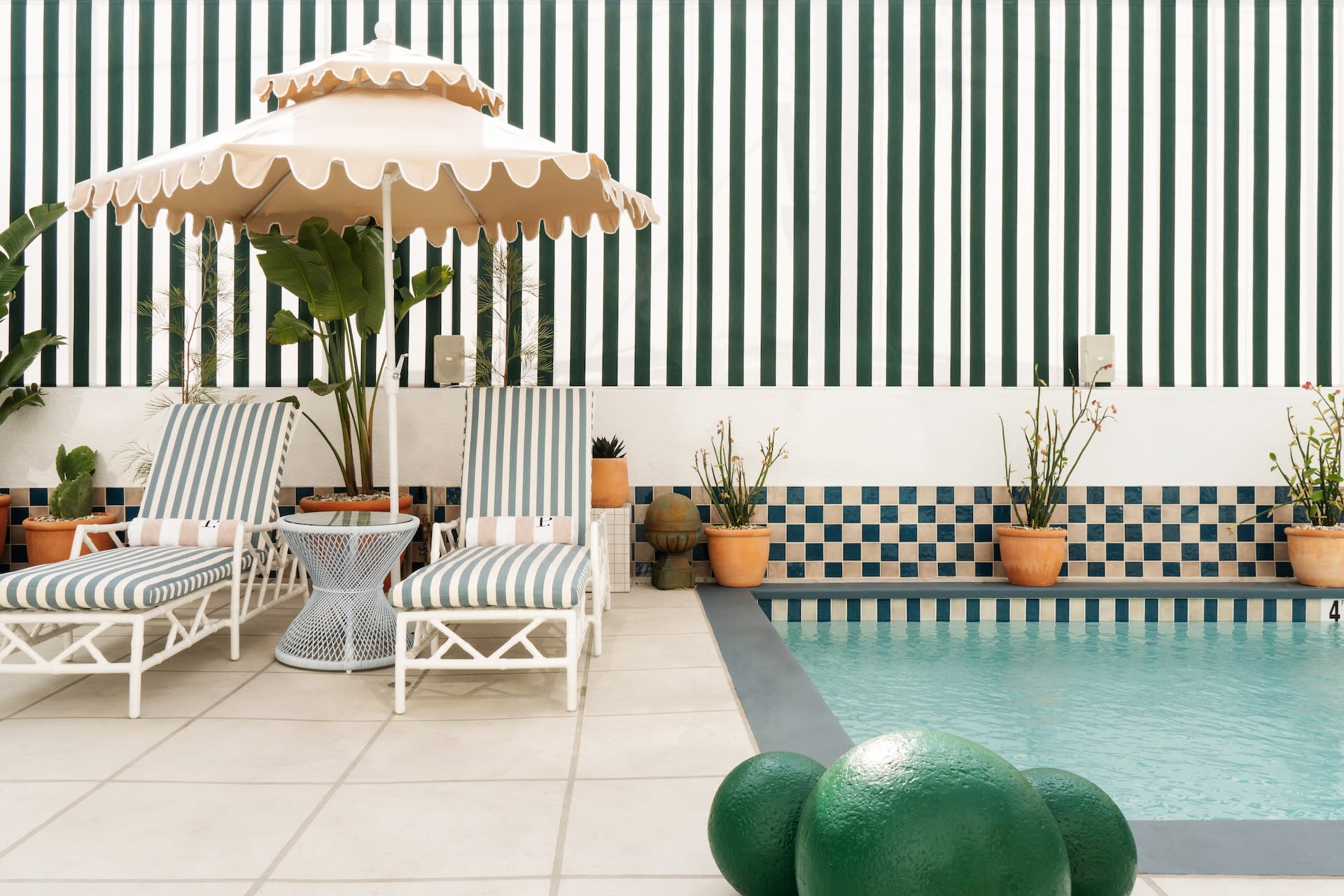 Chic poolside scene with striped lounge chairs, a scalloped umbrella, potted plants, and a turquoise pool framed by green-and-white striped walls and tiled accents.
