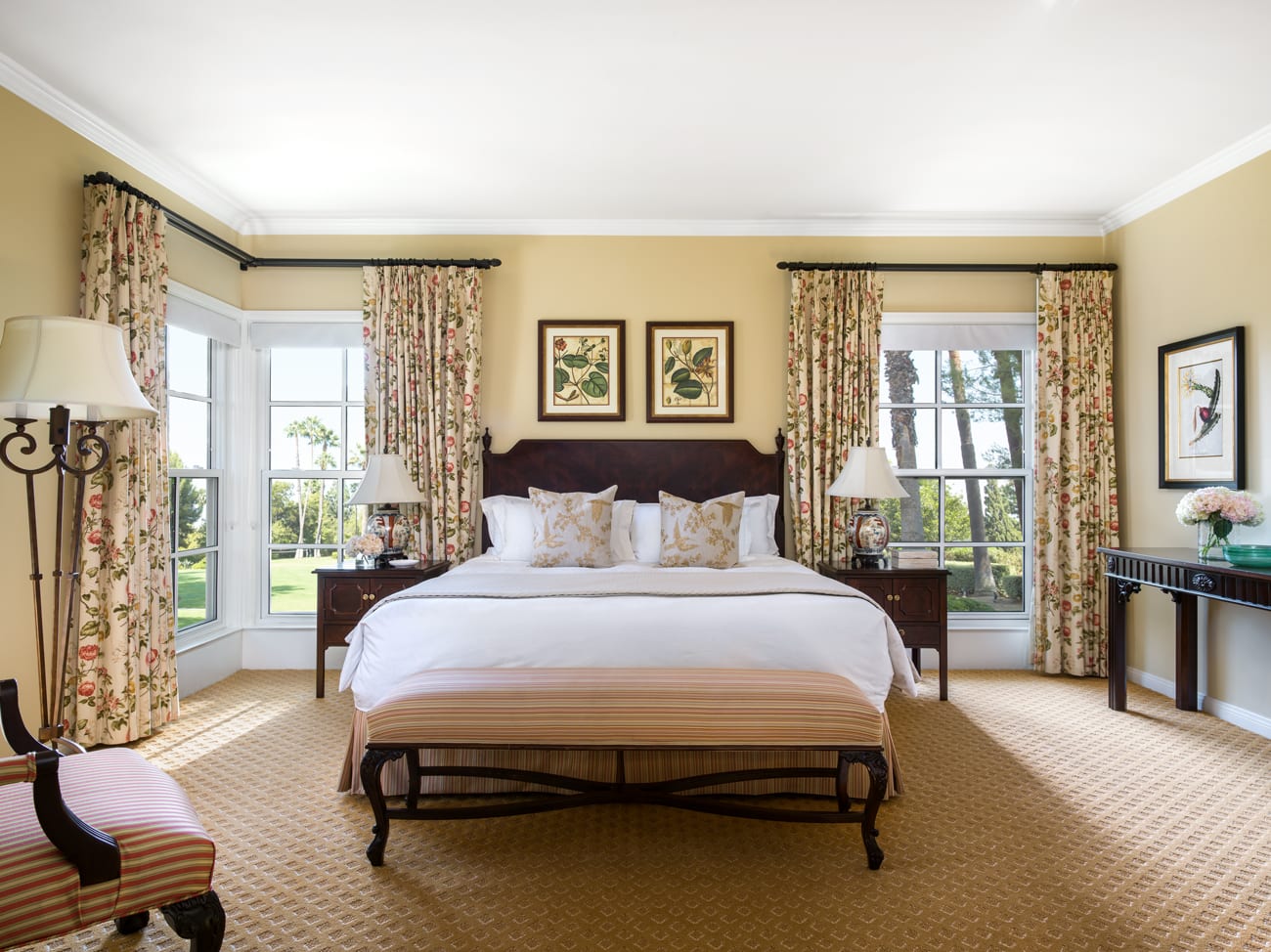 Elegant traditional bedroom with floral curtains, framed botanical art, and a large wooden bed dressed in white linens and gold-accented pillows overlooking a garden view.