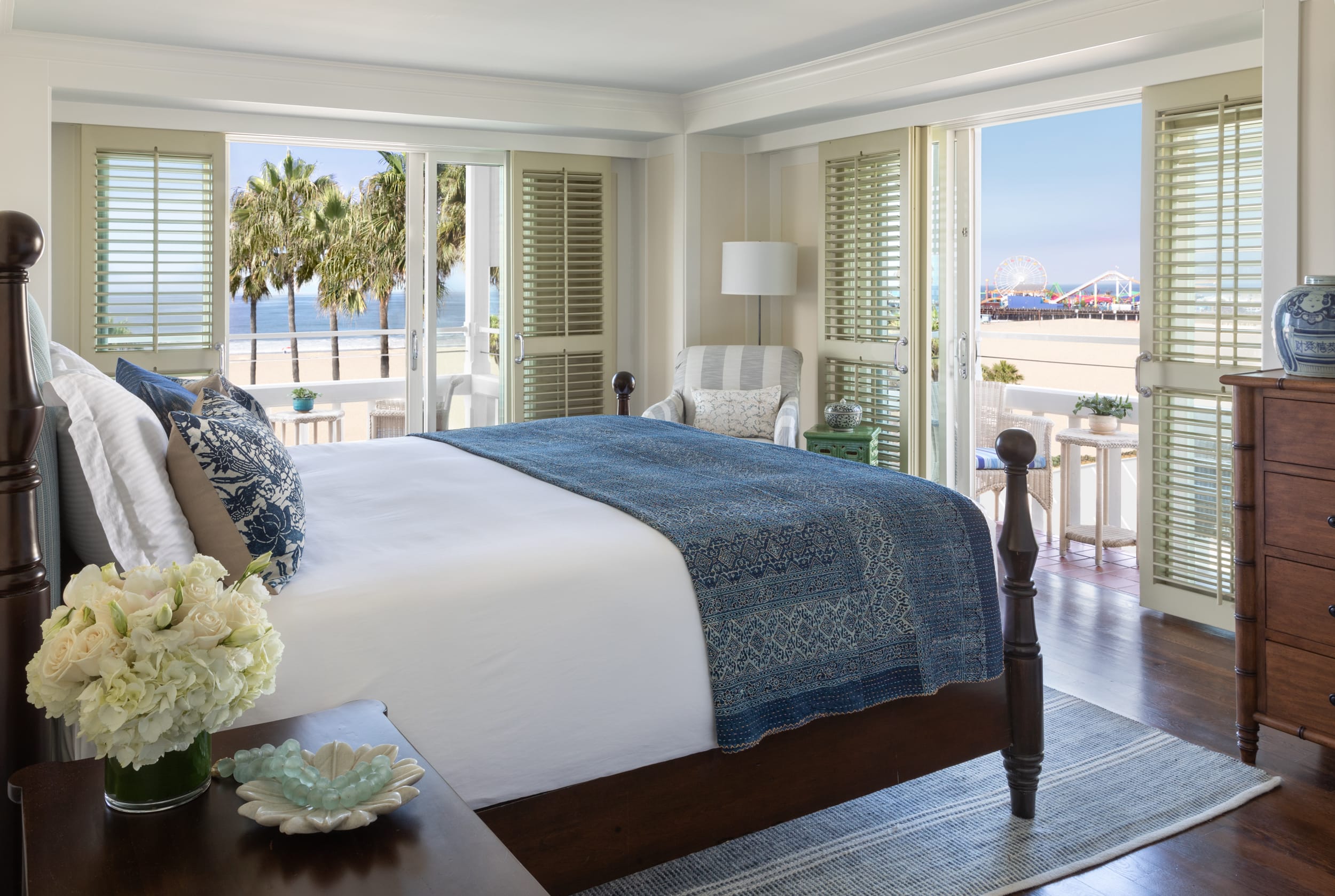 Bright coastal bedroom with ocean views, featuring a four-poster bed with blue accents, large windows opening to a balcony, and Santa Monica Pier visible in the distance.