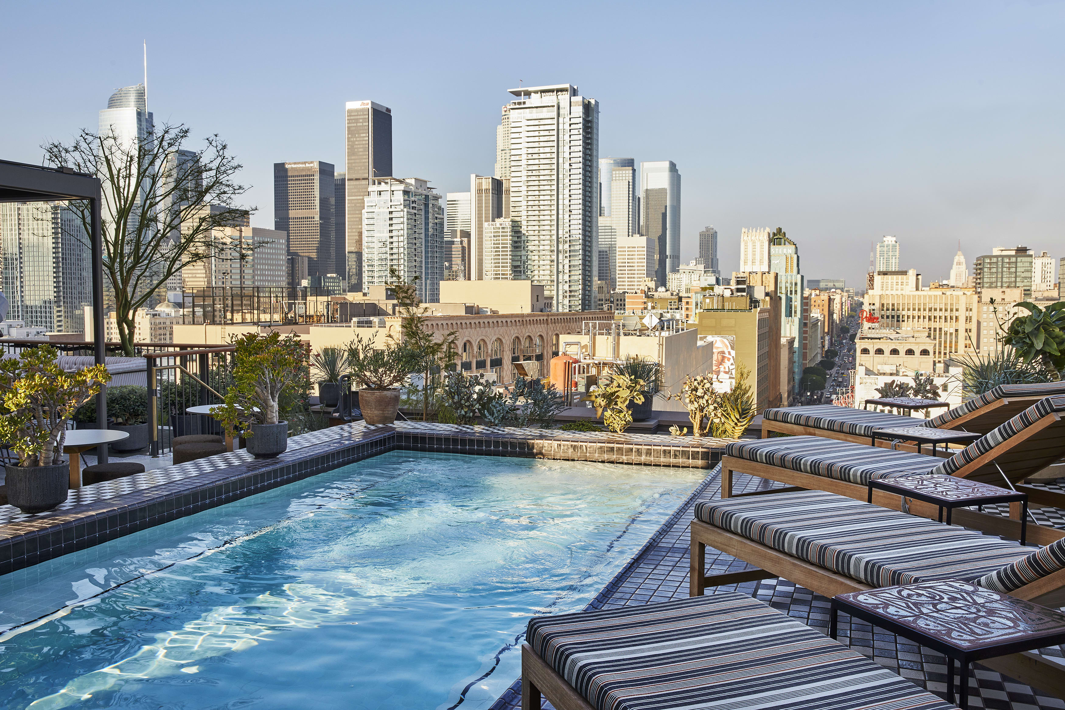 Rooftop pool with striped lounge chairs and potted plants overlooking the Los Angeles skyline under a clear afternoon sky.