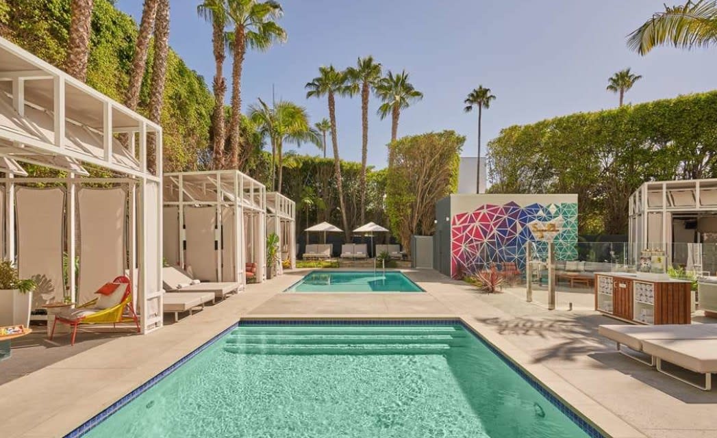 Modern outdoor pool area at a hotel with white cabanas, palm trees, and a colorful geometric mural under a bright California sky.