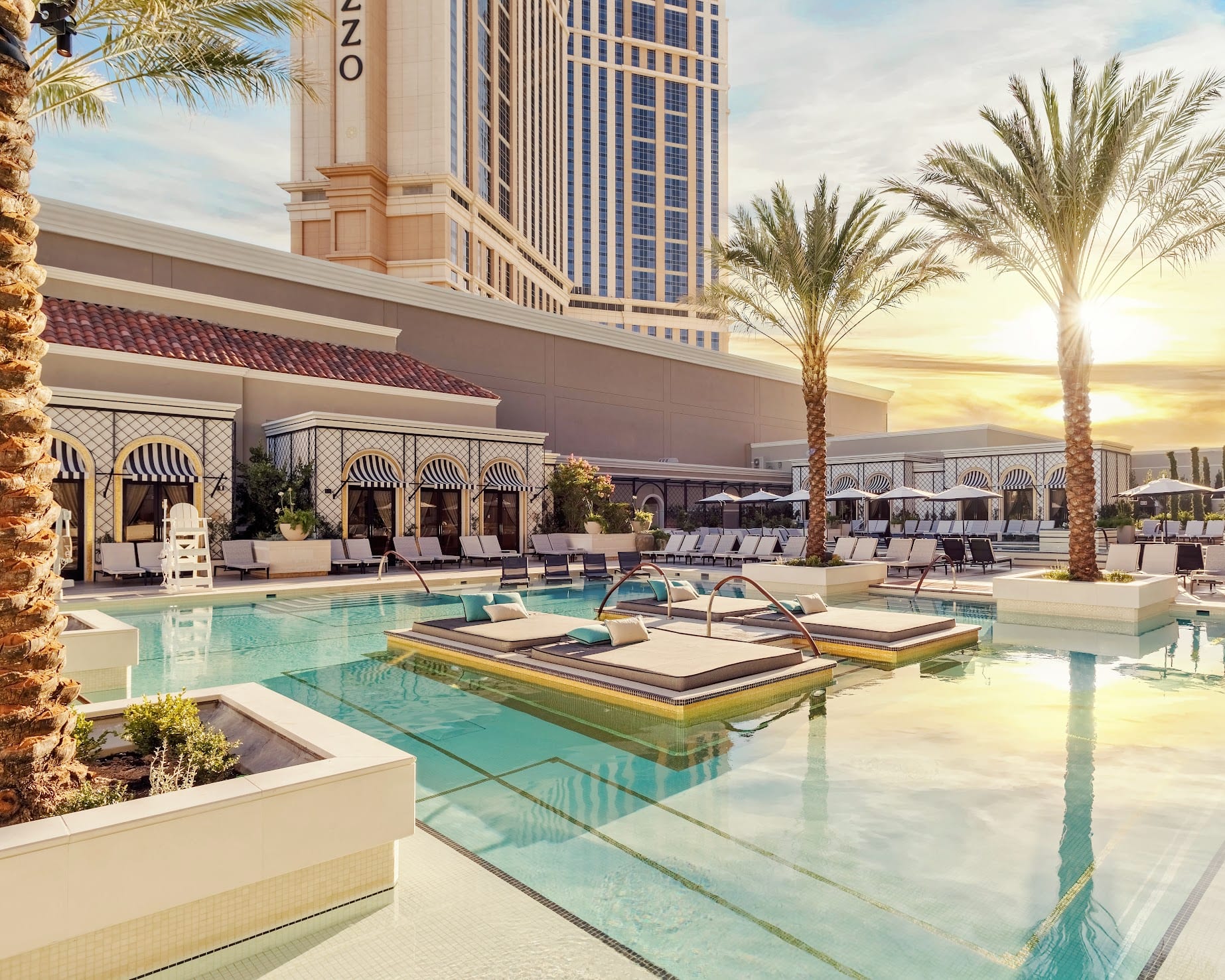 a resort pool with lounge chairs and palm trees, a towering hotel and sun rising in the background