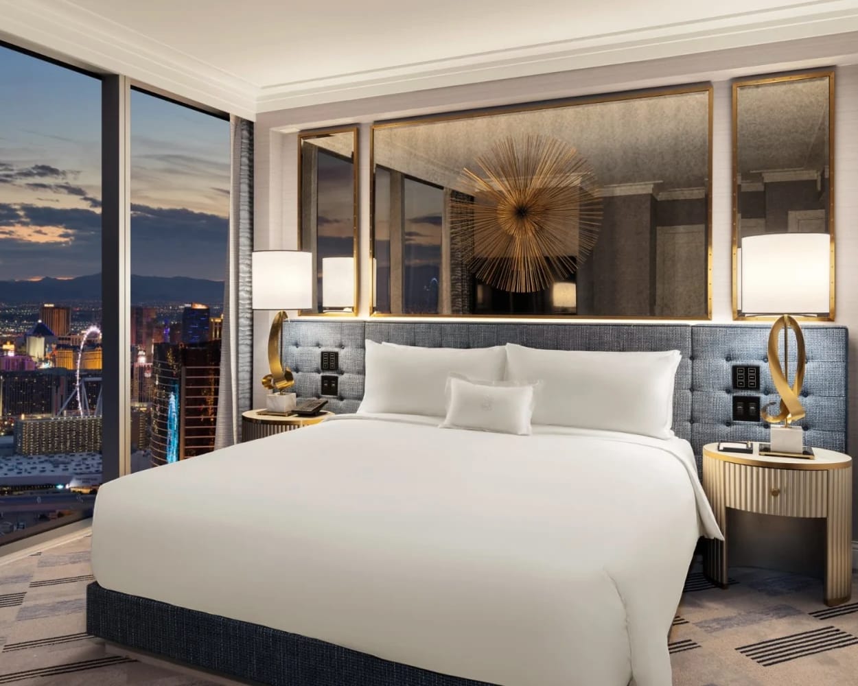 A modern hotel room with white bed linens, gold accents, mirror headboard, and city lights outside the window