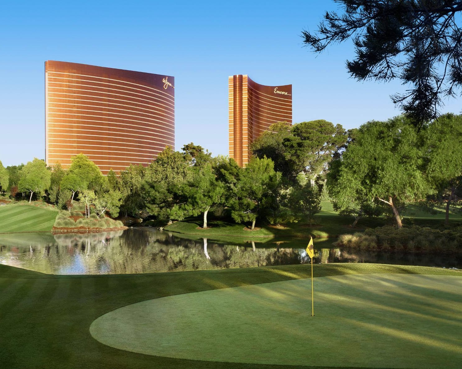 a golf course with lake, trees, and two bronze skyscrapers behind