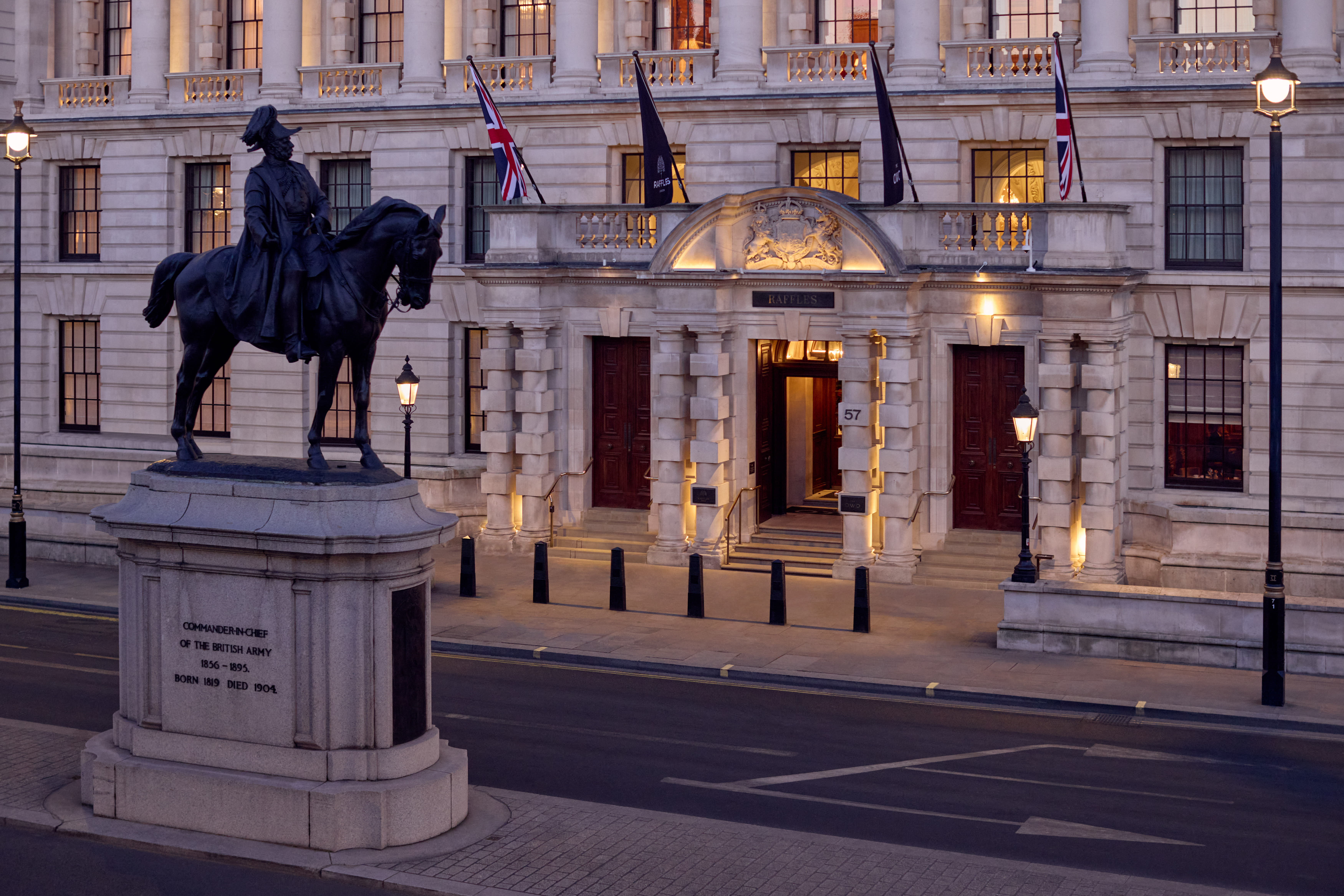Grand exterior of a neoclassical building with Union Jack flags and warm lighting, featuring a bronze equestrian statue in the foreground.