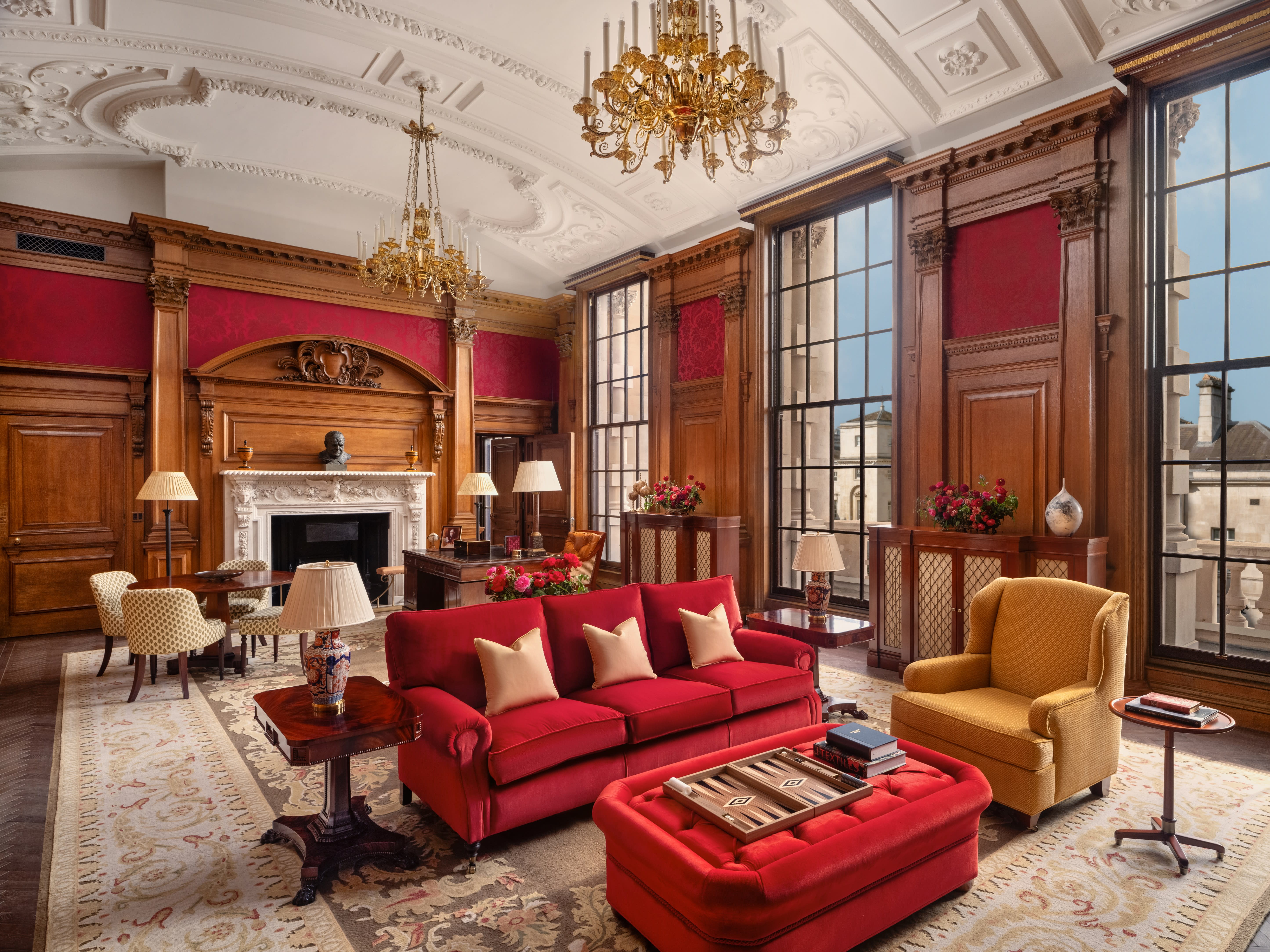 Elegant lounge with red and gold furniture, tall windows, ornate chandeliers, and a carved fireplace framed by wood paneling and classical details.