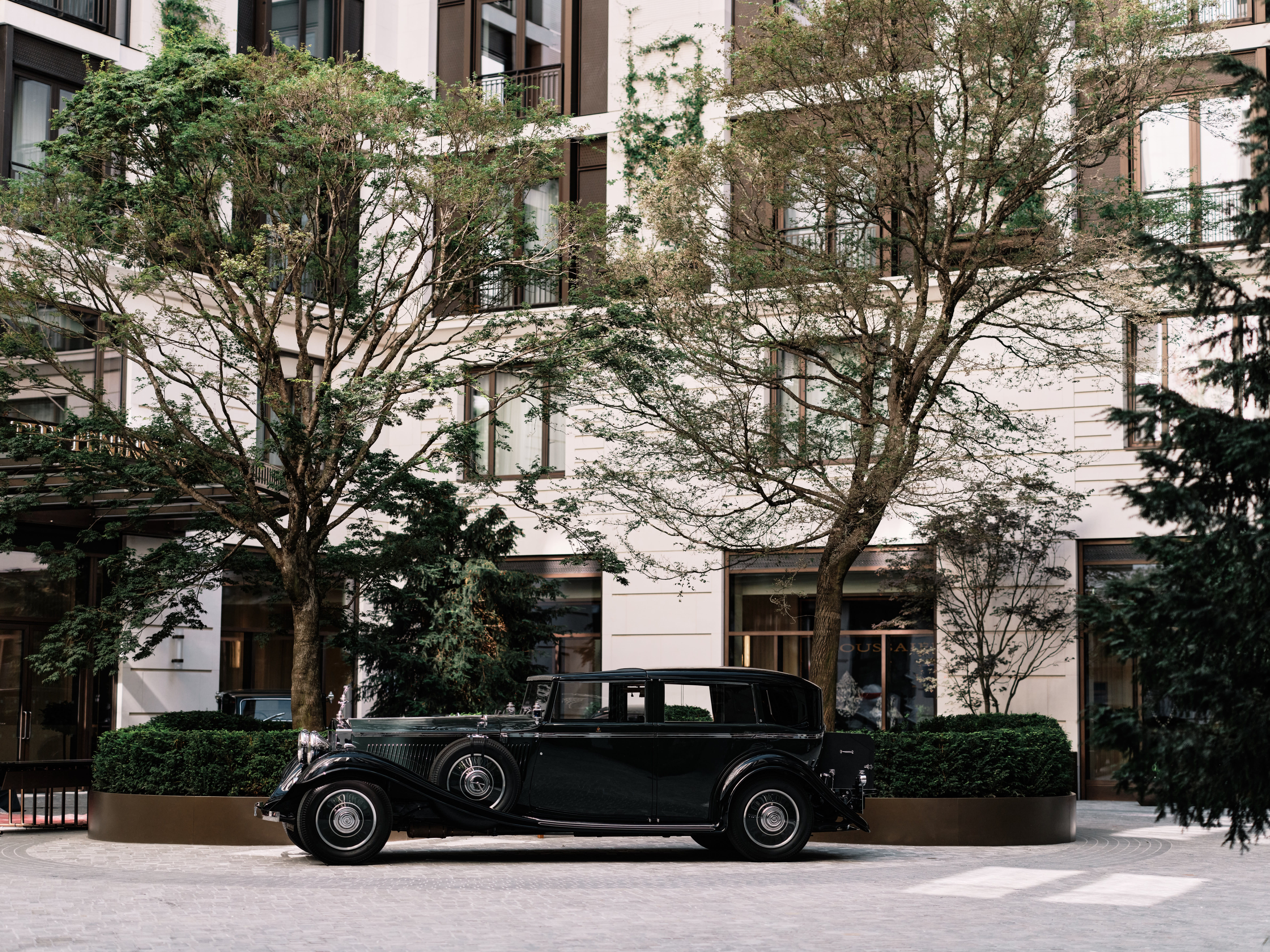 Vintage black Rolls-Royce parked in a tree-lined courtyard outside a modern luxury hotel building.