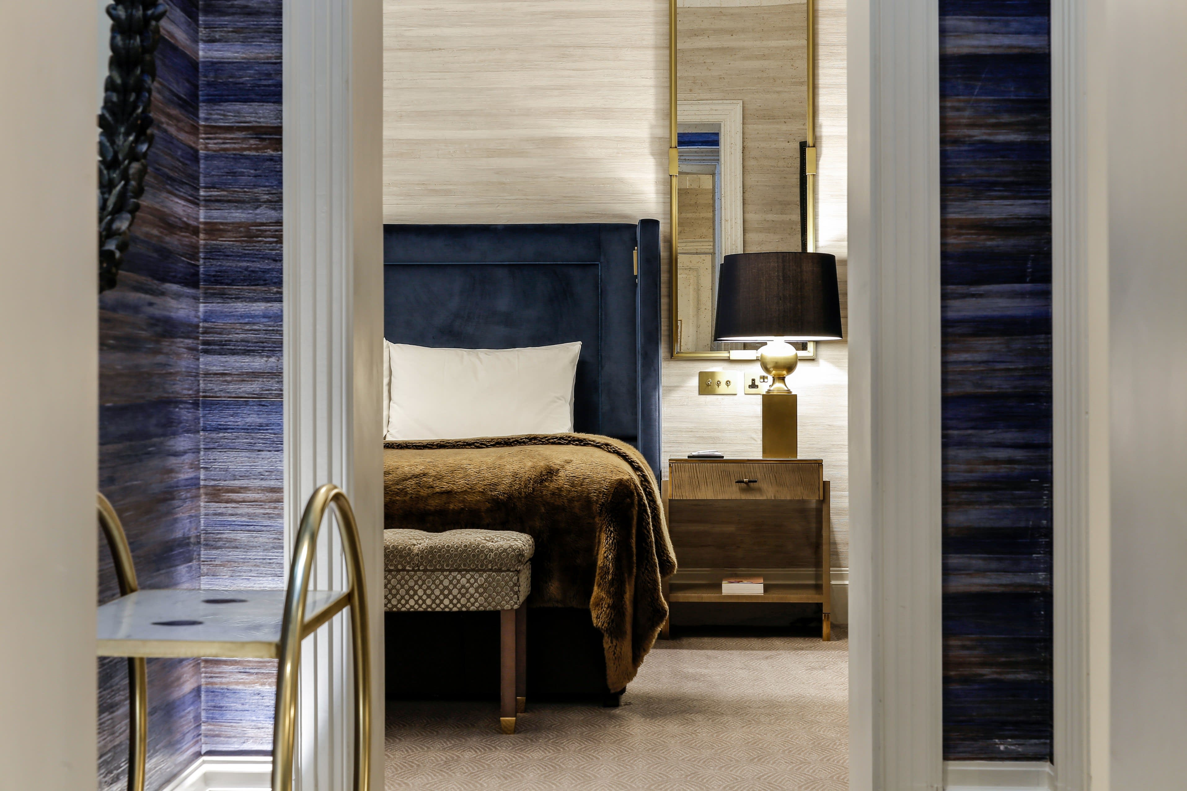 Elegant hotel bedroom with a navy velvet headboard, gold accents, a bedside lamp, and rich textures in blue and brown tones.