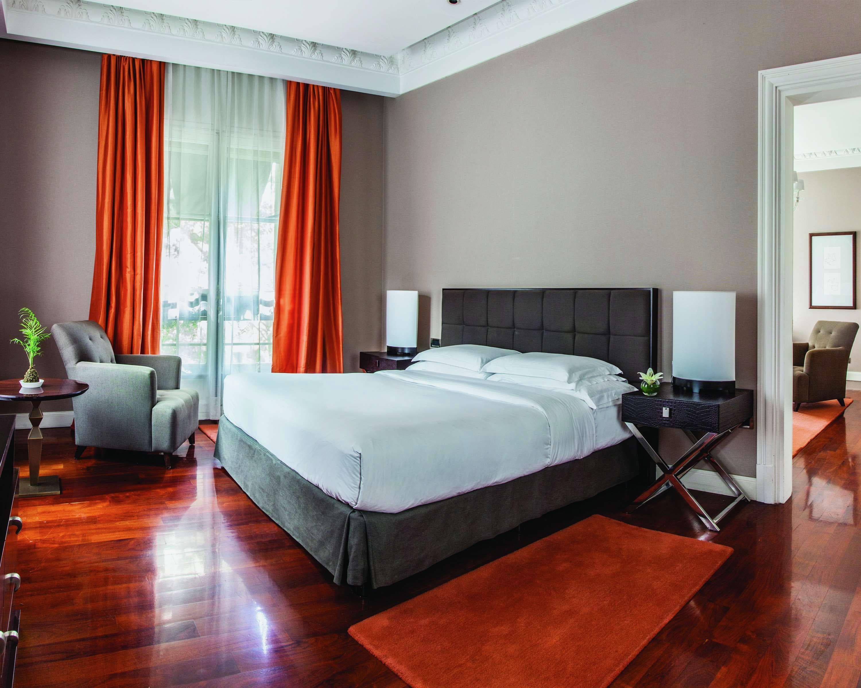 A hotel room with red wood floors and orange curtains, grey fabric chair and bed, and brown walls