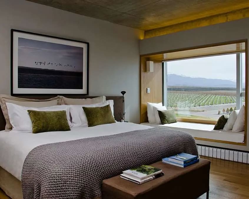 hotel room with white linen bed, green accent pillows, and grey blanket, plus wood floors and window seat overlooking vineyards