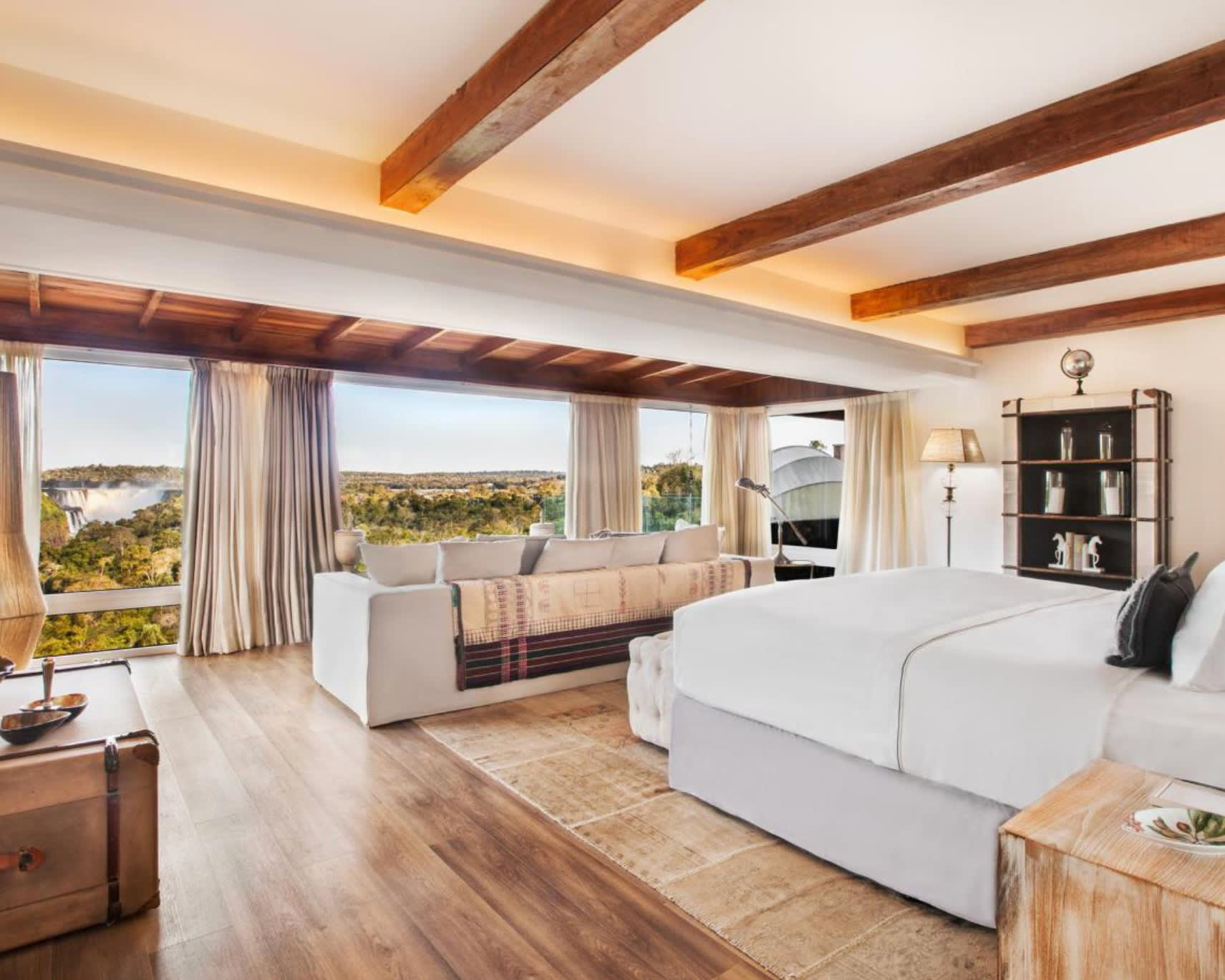 modern hotel room with white bed, walls, and furniture, wood beam ceiling and wood floors, with jungle landscape and waterfalls out the windows