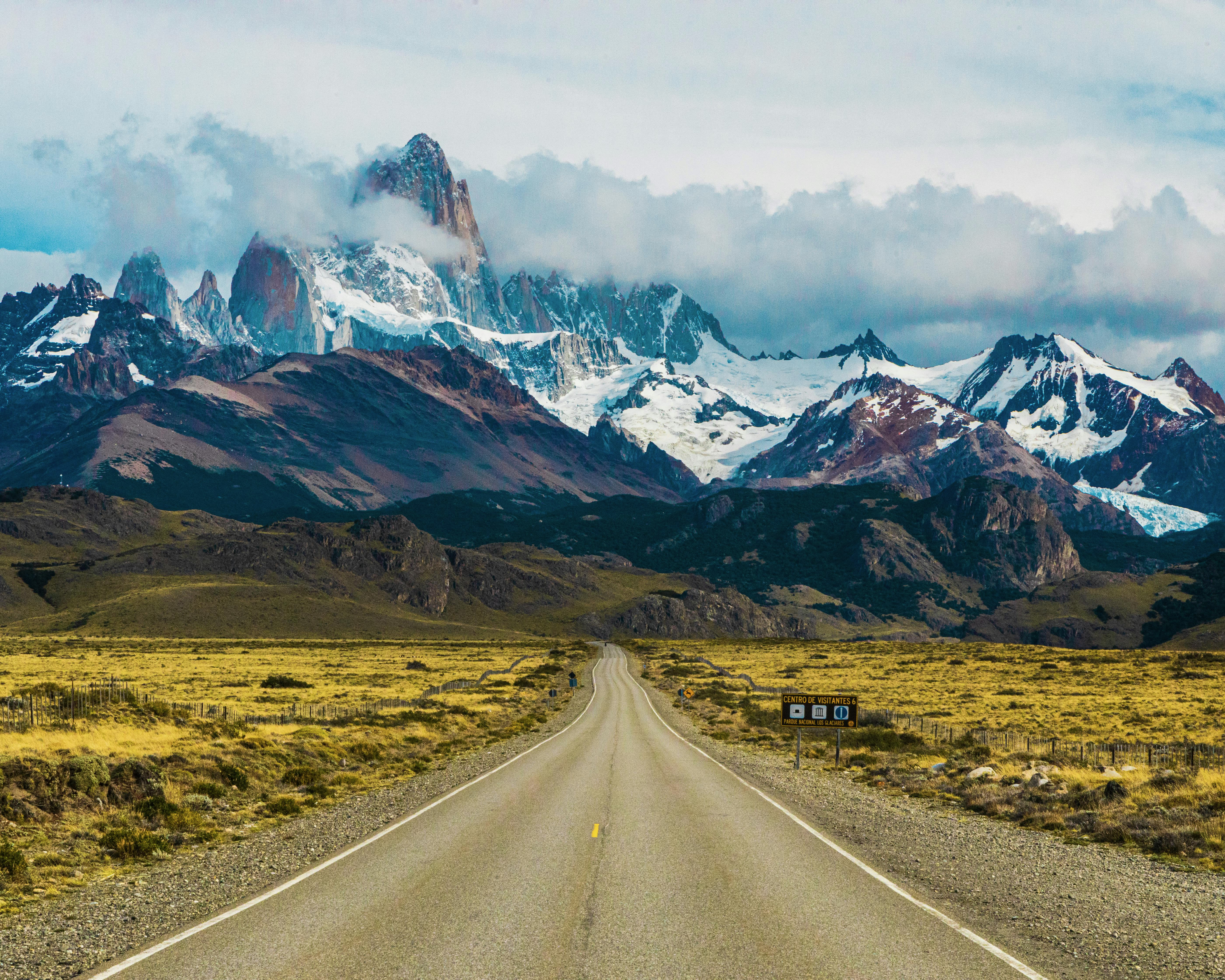 a road through a green valey with tall brown snow-covered mountains and clouds during day
