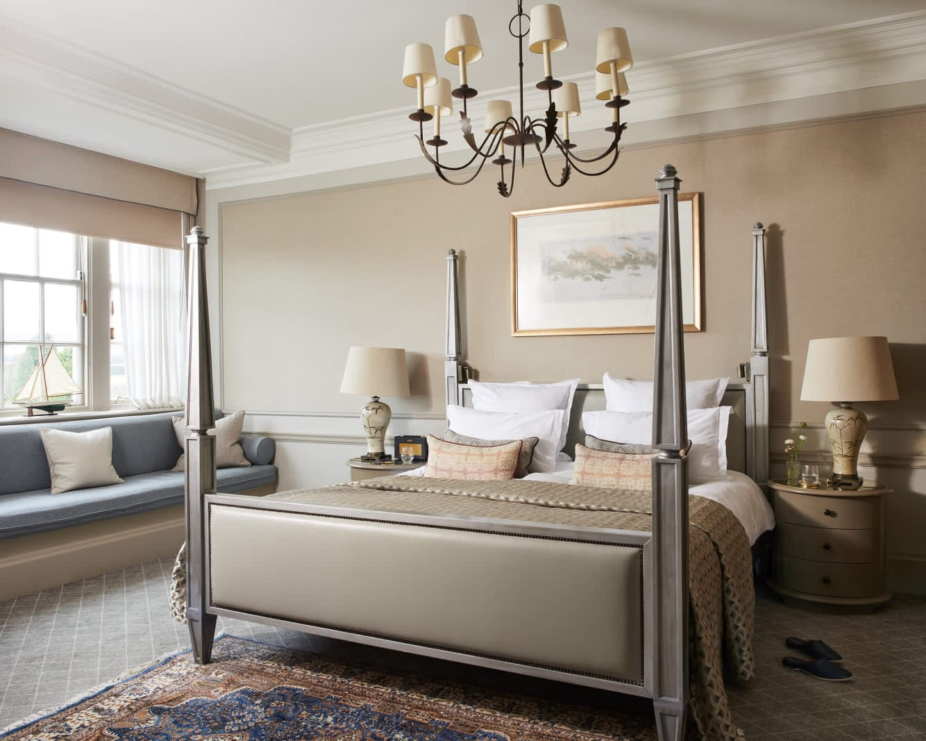 classic hotel room with beige four poster bed, beige walls, art, and chandelier over the bed