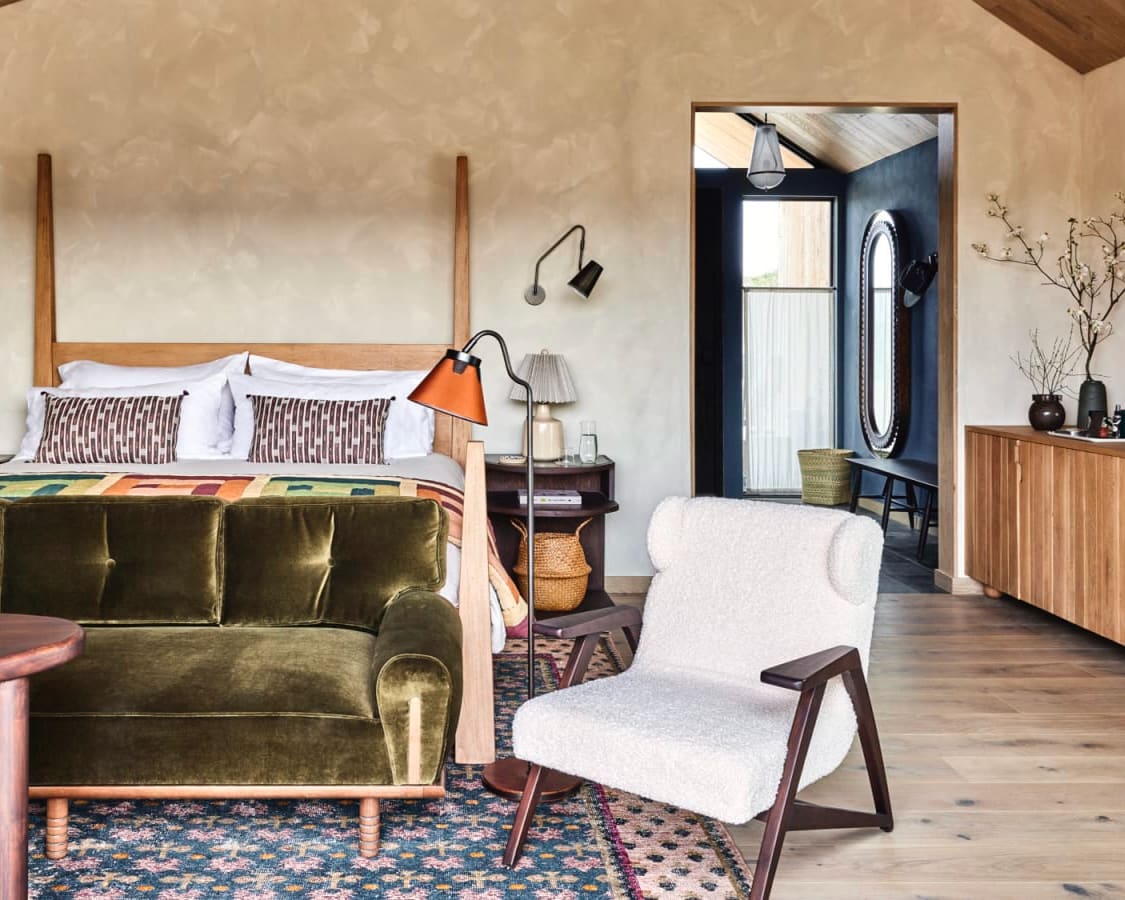rustic modern hotel room with tan walls, green sofe, white chair, and four poster bed