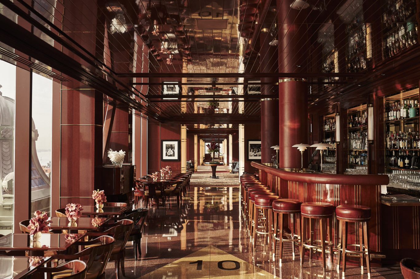 Elegant hotel bar and lounge with rich mahogany interiors, red leather stools, polished floors, and large windows offering city and sea views.