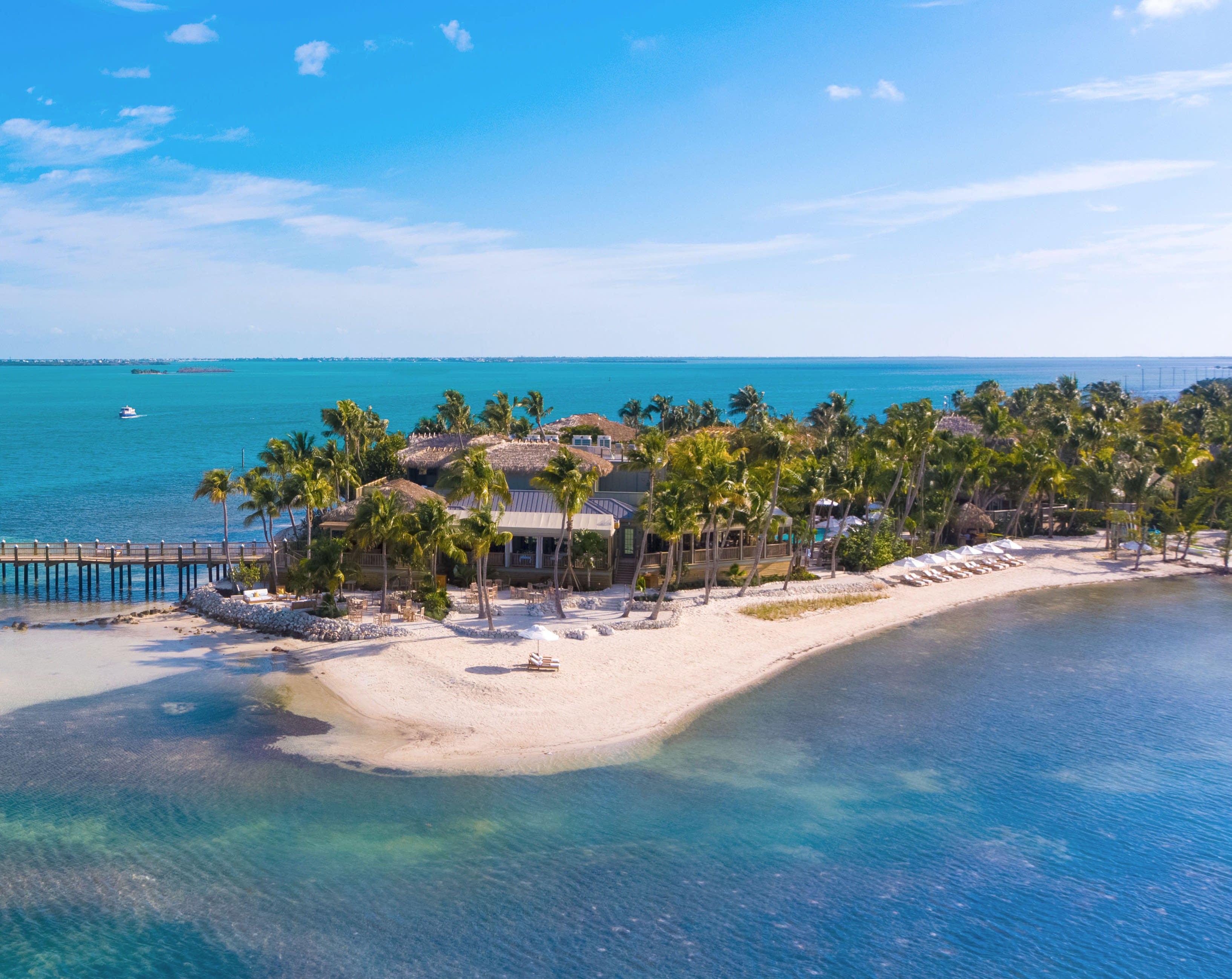 Aerial view of a tropical island resort with white-sand beaches, turquoise waters, palm trees, and a long wooden dock extending into the sea.