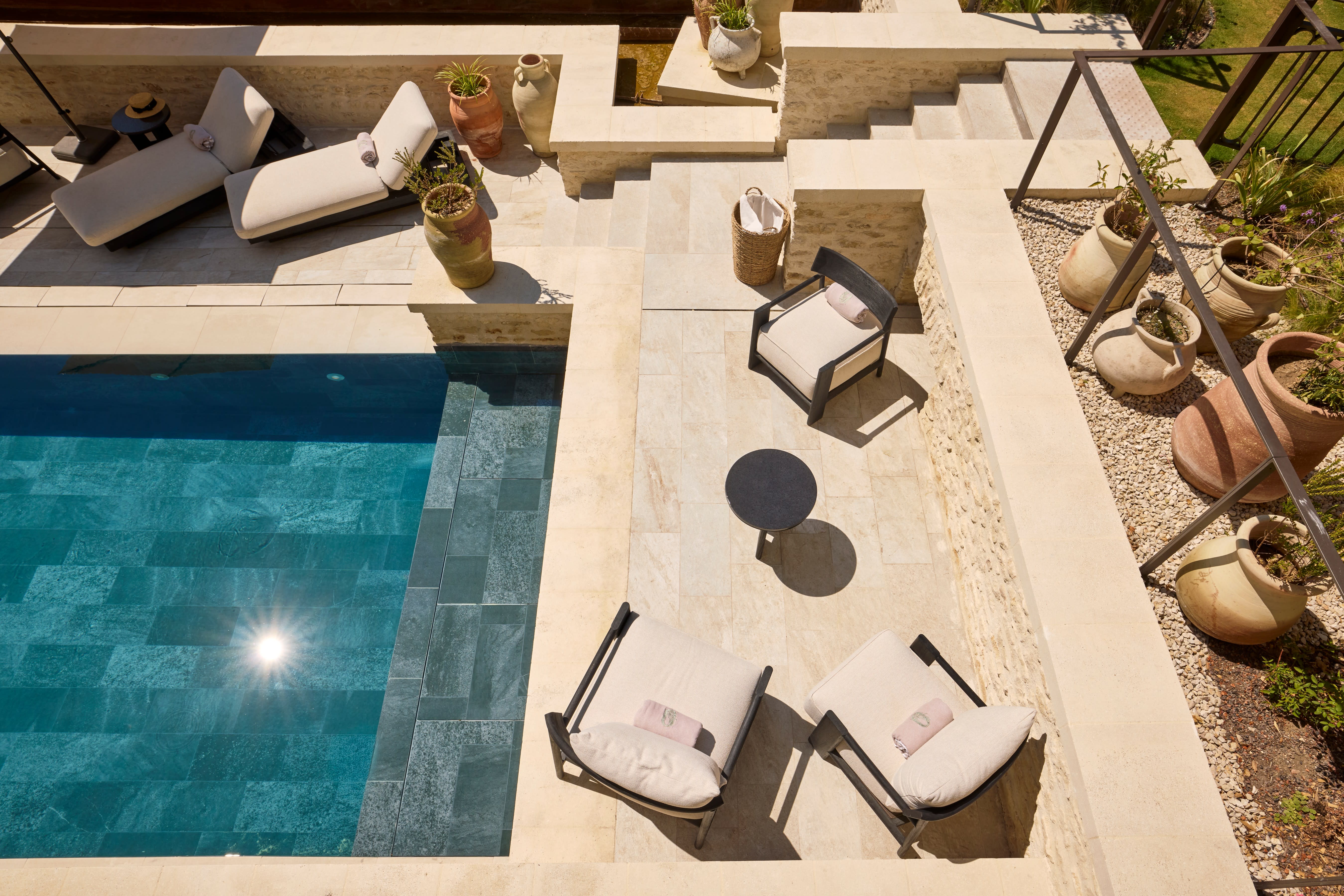 Sunlit stone terrace with a turquoise-tiled pool, cushioned lounge chairs, terracotta pots, and a cozy seating area creating a Mediterranean-style retreat.