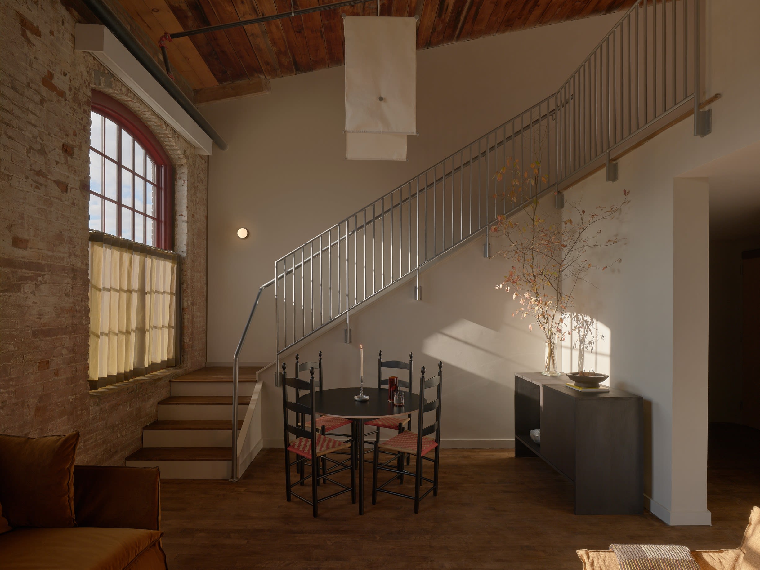 Warm industrial-style loft with exposed brick walls, wooden ceiling, and a metal staircase leading to an upper level, featuring minimalist furniture and natural light.