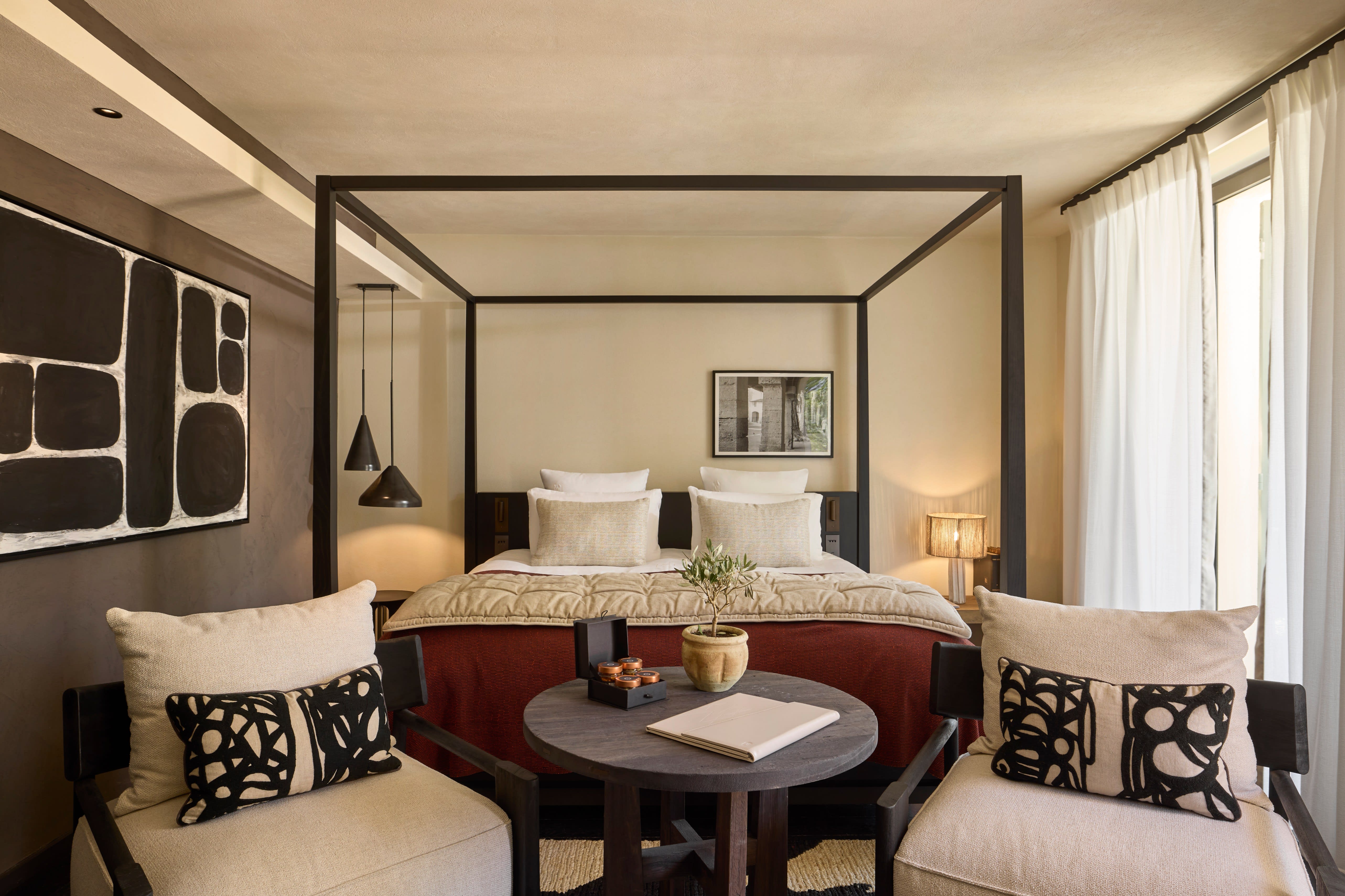 Elegant bedroom with a modern four-poster bed, neutral tones, abstract black artwork, and cozy seating with patterned cushions and soft natural light.
