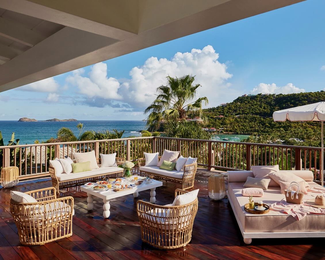 an outdoor rooftop patio with plush furniture overlooking a tropical ocean and hillside during day