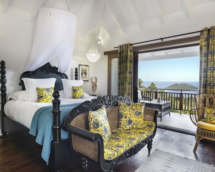 an eclectic hotel room with black four poster bed, teal banket, yellow and green accent pillows, chairs, and drapers overlooking the ocean
