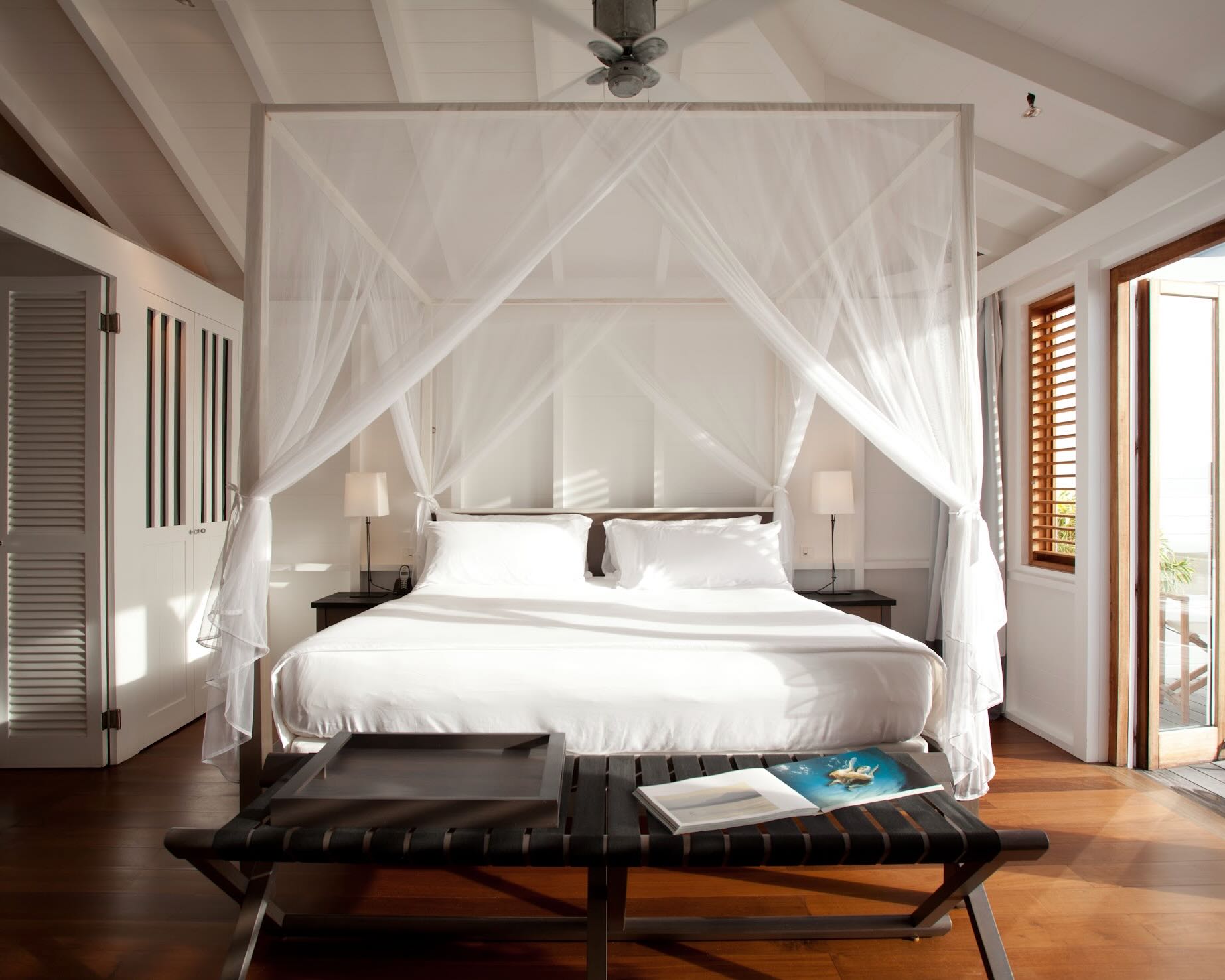 four poster bed with shear curtains, white linens, black bench at the foot of the bed, and white wood beam ceiling