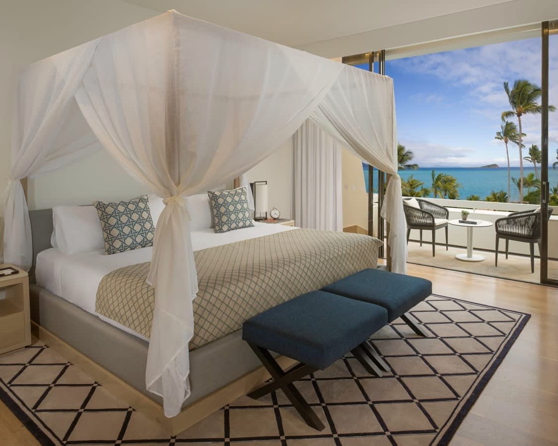 a four poster bed with white curtains, teal ottomans at the foot of the bed, and a palm tree lined ocean view outside