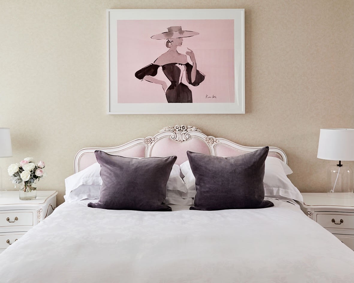 pink tufted headboard of a bed with grey pillows, white side tables with lamp and flowers, and a pink framed artwork on the wall above the bed