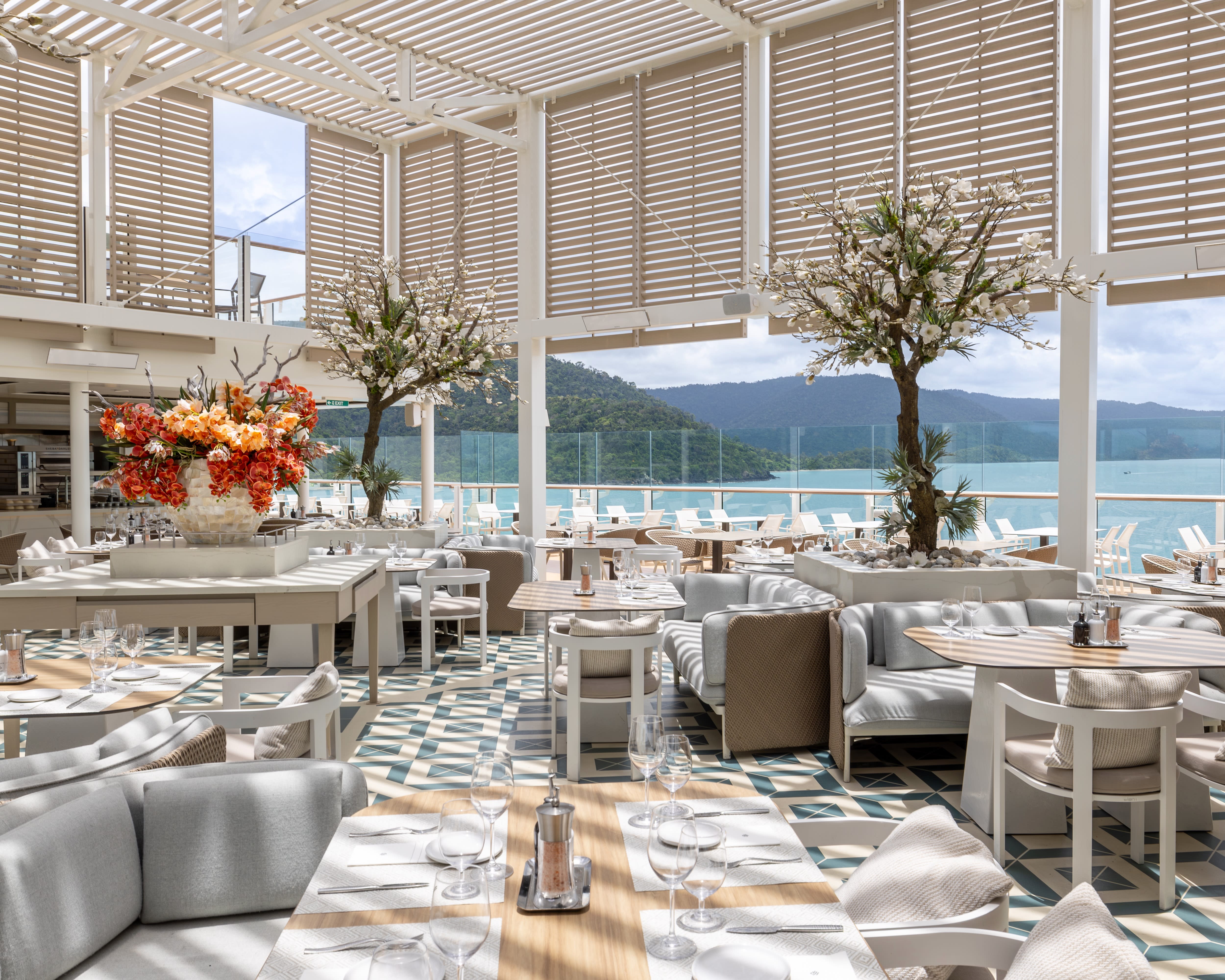 modern dining area with white tables and chairs, grey patterned flooring, flowers and vaulted ceilings with an island outside