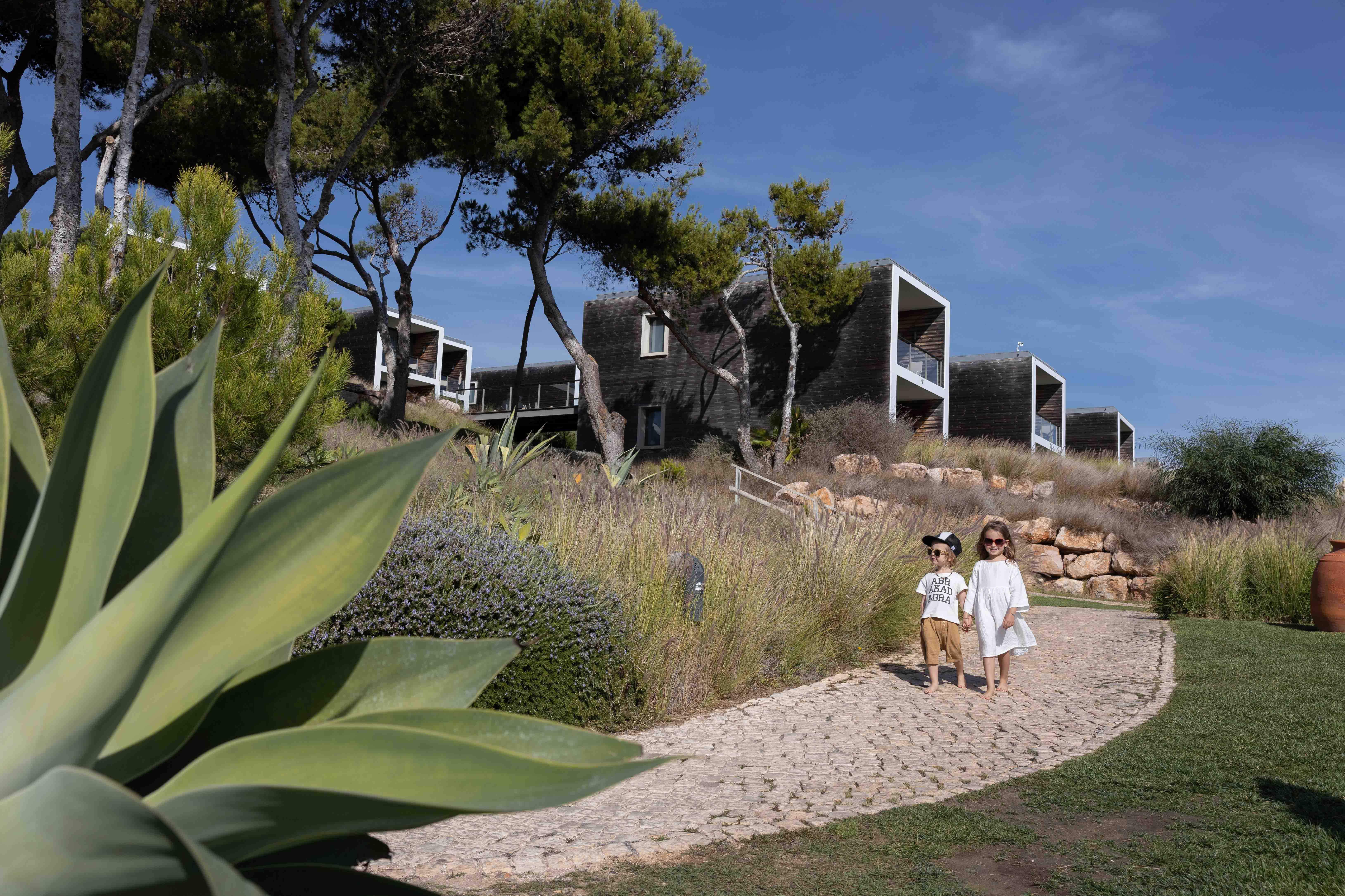 Two children walking on a stone path surrounded by greenery and modern villa-style buildings at a coastal resort with blue skies above.