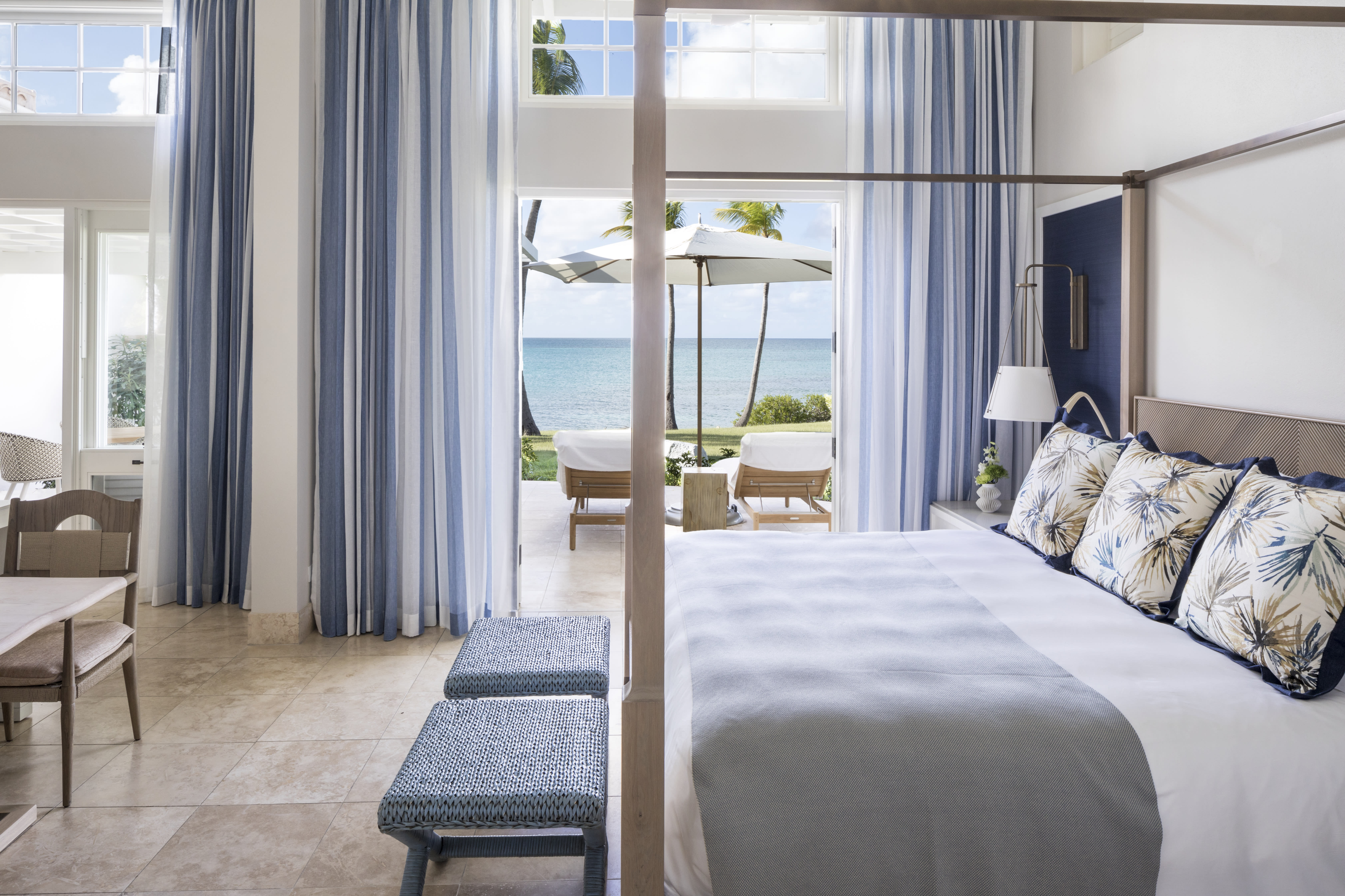 A bright, coastal-inspired bedroom with blue-and-white striped curtains, a four-poster bed, and patterned pillows, opening onto a terrace with sun loungers, an umbrella, and views of palm trees and the turquoise ocean.