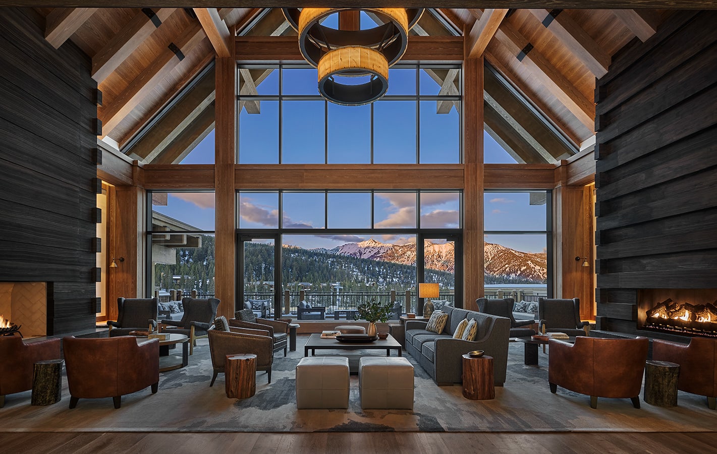 A spacious mountain lodge lounge with high vaulted wooden ceilings, large floor-to-ceiling windows framing snow-covered peaks, cozy seating areas with leather and fabric armchairs, and a glowing fireplace creating a warm, welcoming atmosphere.