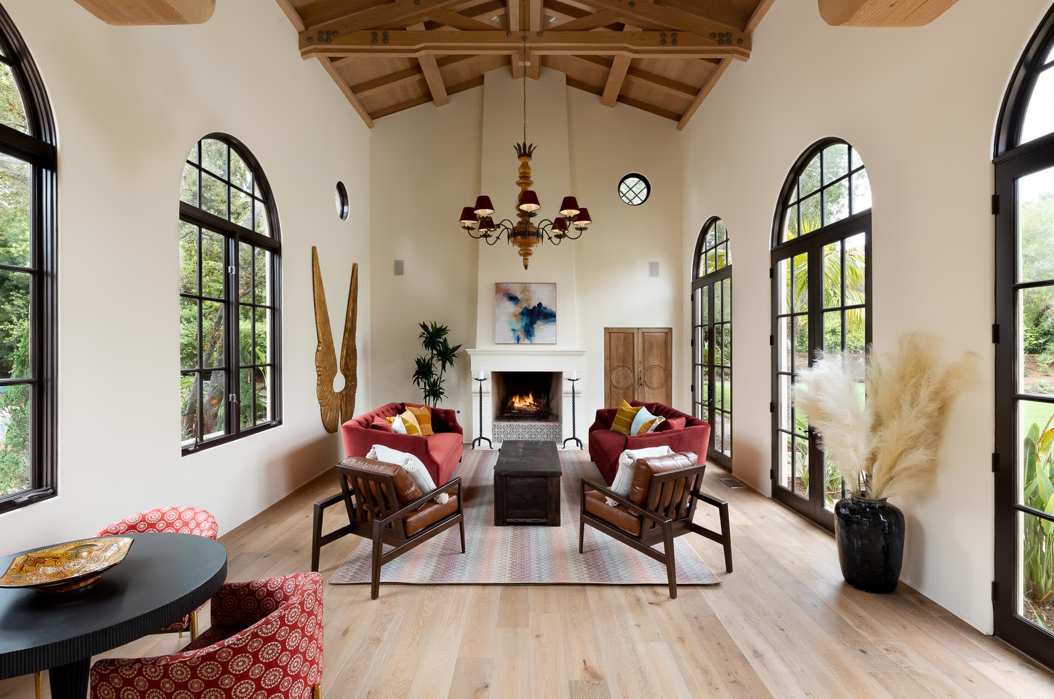 A bright, Mediterranean-style living room with tall arched windows, exposed wood-beam ceilings, and a central fireplace..