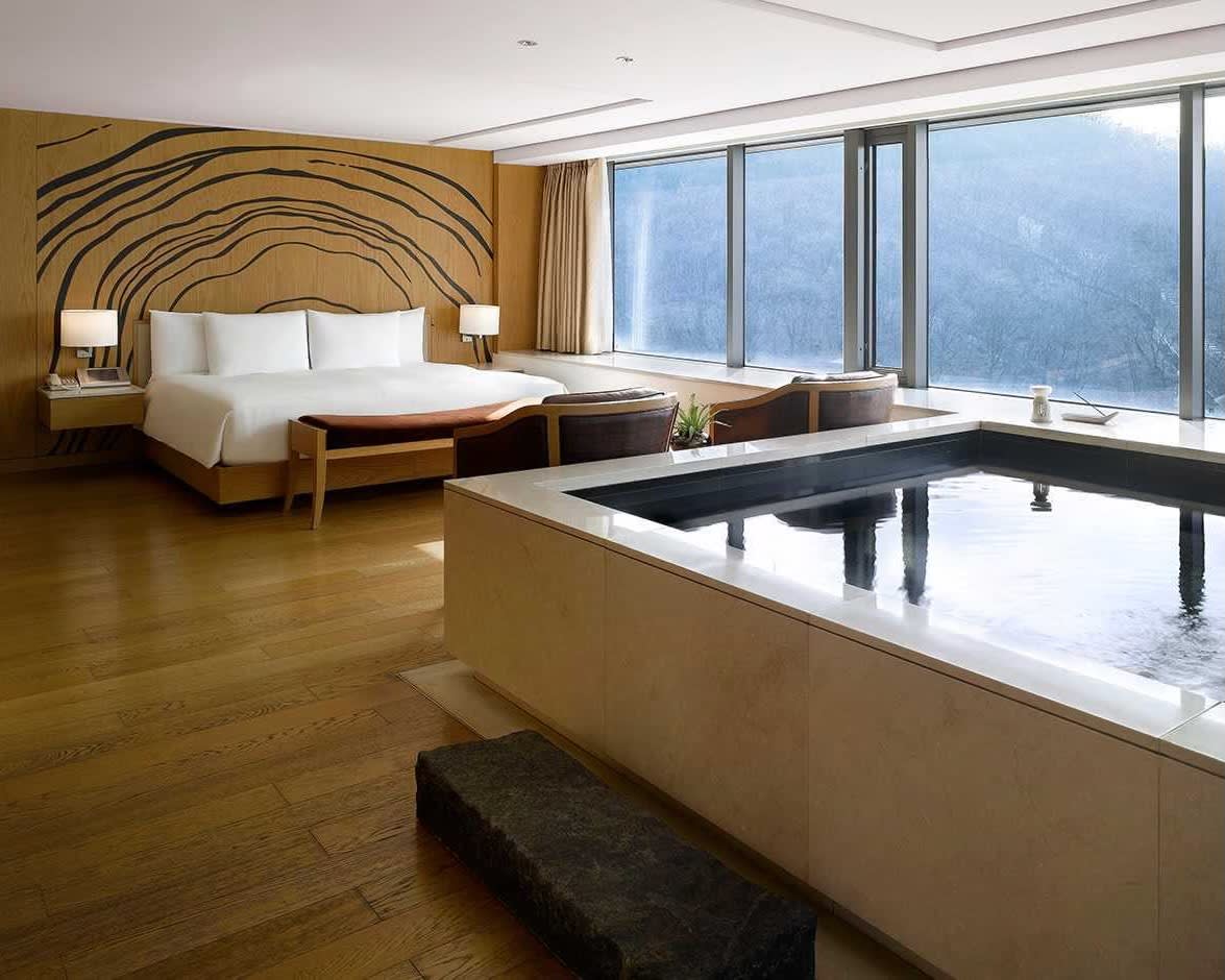 modern, minimalist hotel room with white sheets on the bed, tan and black patterened wall, tan wood floors, and jacuzzi pool in the room