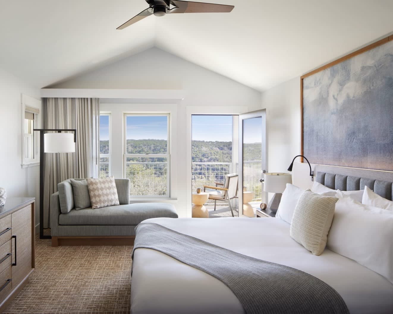 room with white bed, blue painting above the bed, and chaise lounge under windows overlooking a forest landscape