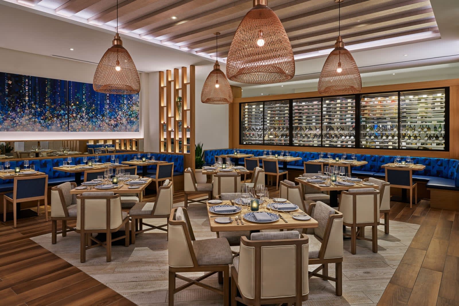 modern restaurant with tan chairs and tables, wood floors, blue accents and artwork, below wood chandeliers
